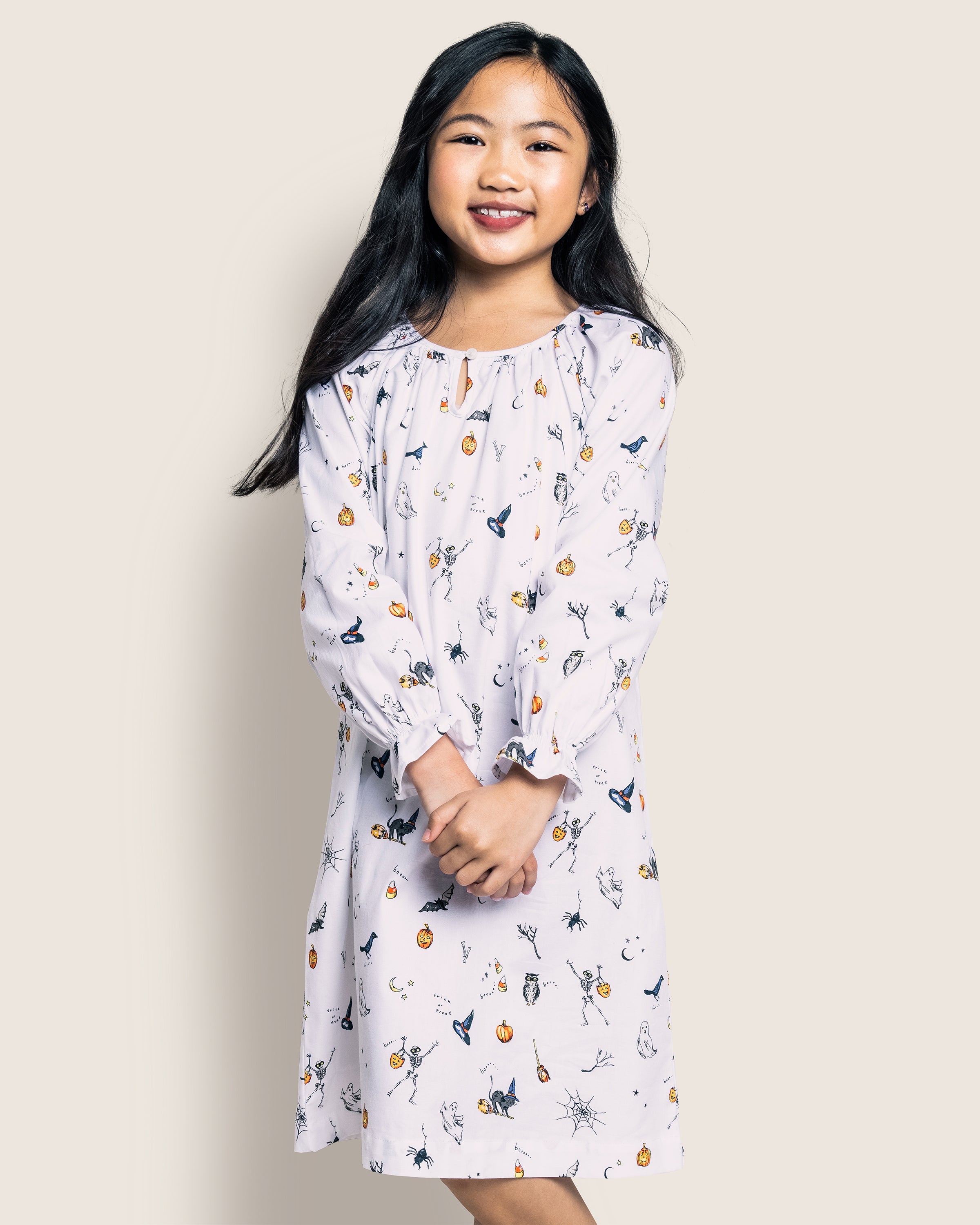 A girl with long black hair wears the Petite Plume Girls Twill Delphine Nightgown in Trick or Treat, featuring whimsical Halloween-themed icons. Standing against a neutral background, her enchanting look is enhanced by her light-colored dress and clasped hands.