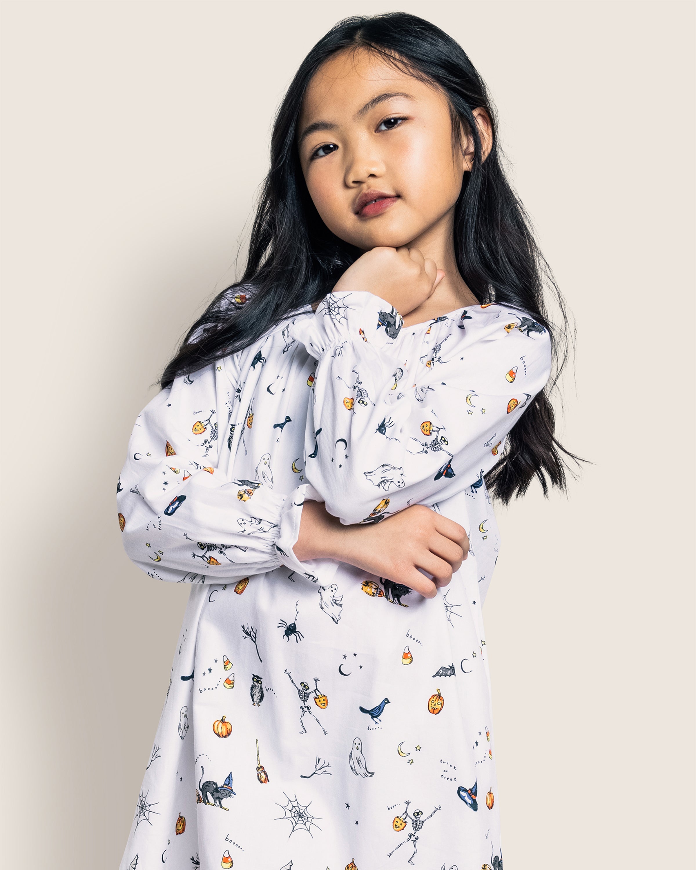 A young girl poses against a neutral background in Petite Plumes Girls Twill Delphine Nightgown in Trick or Treat, featuring colorful Halloween icons. Her long dark hair is loose, and she rests one hand on her chin while the other crosses her waist, exuding confidence.