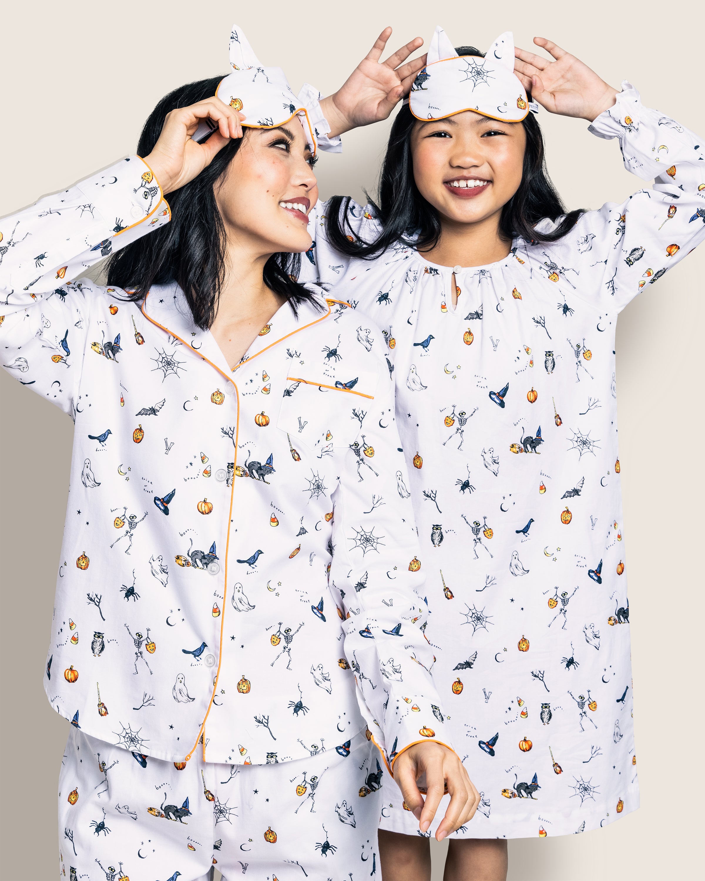 Two people in matching Petite Plume Trick or Treat nightgowns smile under sleep masks with cat ears, adorned with playful Halloween icons like bats, spiders, and pumpkins.
