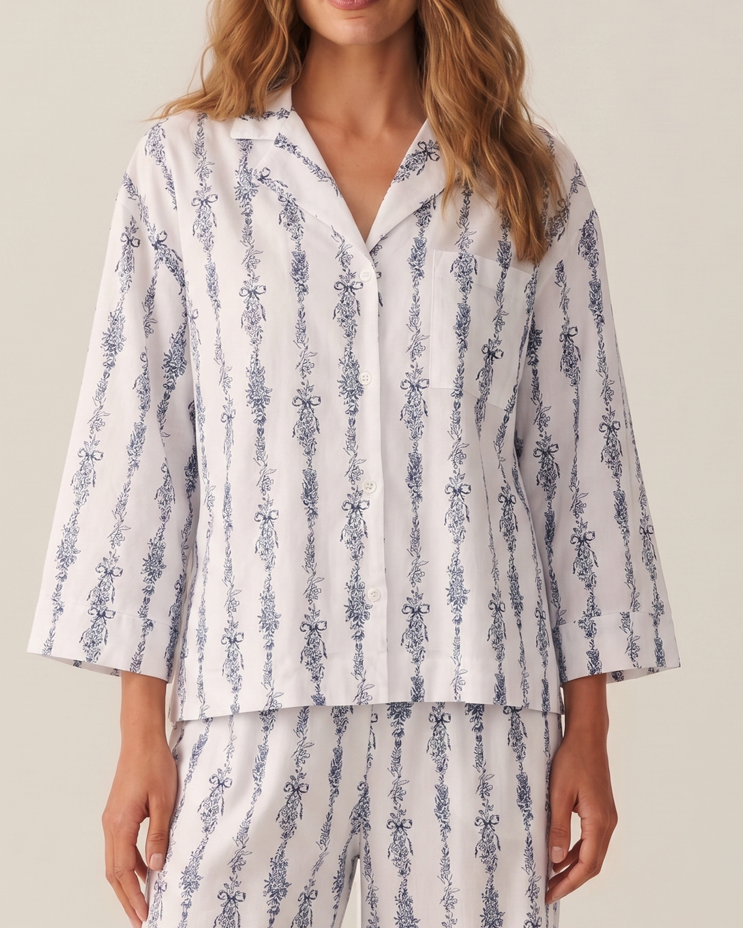 A woman wearing The Olivia Wide Leg Pima Pajama Set in Navy Mayfair Toile, which features a blue and white floral vertical stripe pattern. The pajama top has a collar, buttons, and long sleeves. She is shown from the shoulders to mid-thigh.
