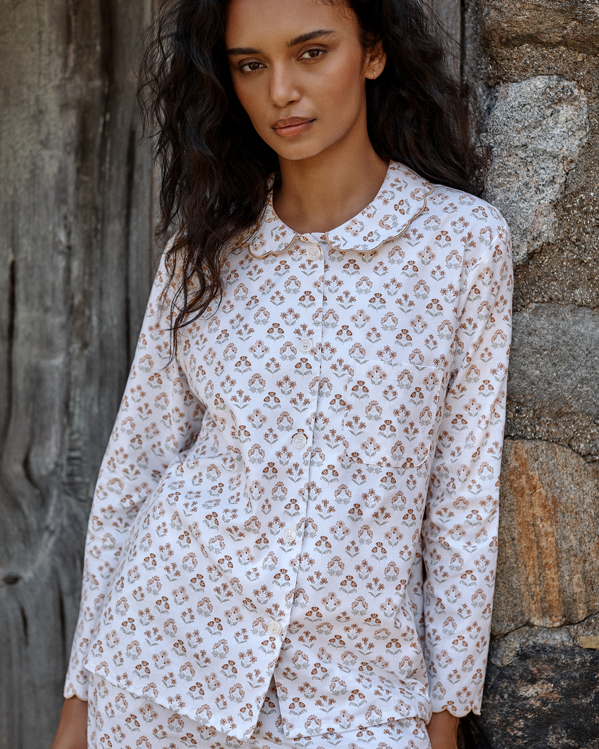 A woman with long dark hair wears the Women’s Twill Colette Pajama Set in Estate Garden, standing in front of a rustic wooden and stone wall and looking at the camera with a relaxed expression.