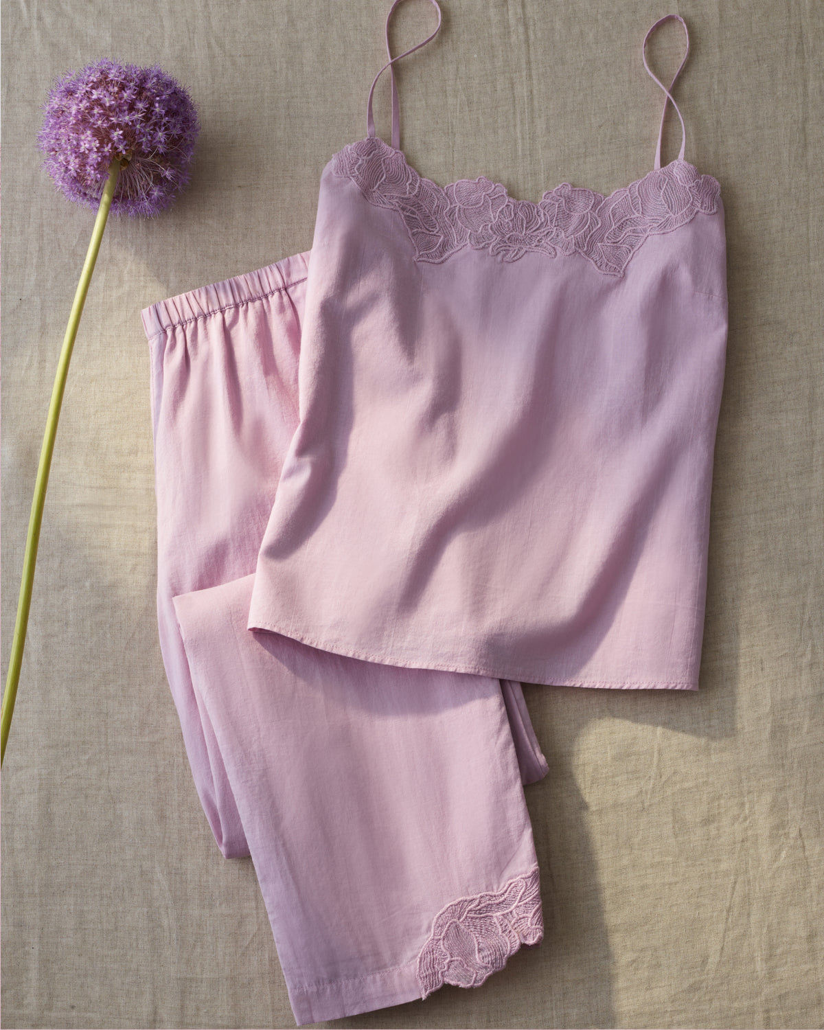 The Women’s Voile Fleur Cami Pant Set in Dusty Rose, featuring delicate lace detailing, is displayed on a beige fabric surface beside a single purple allium flower.