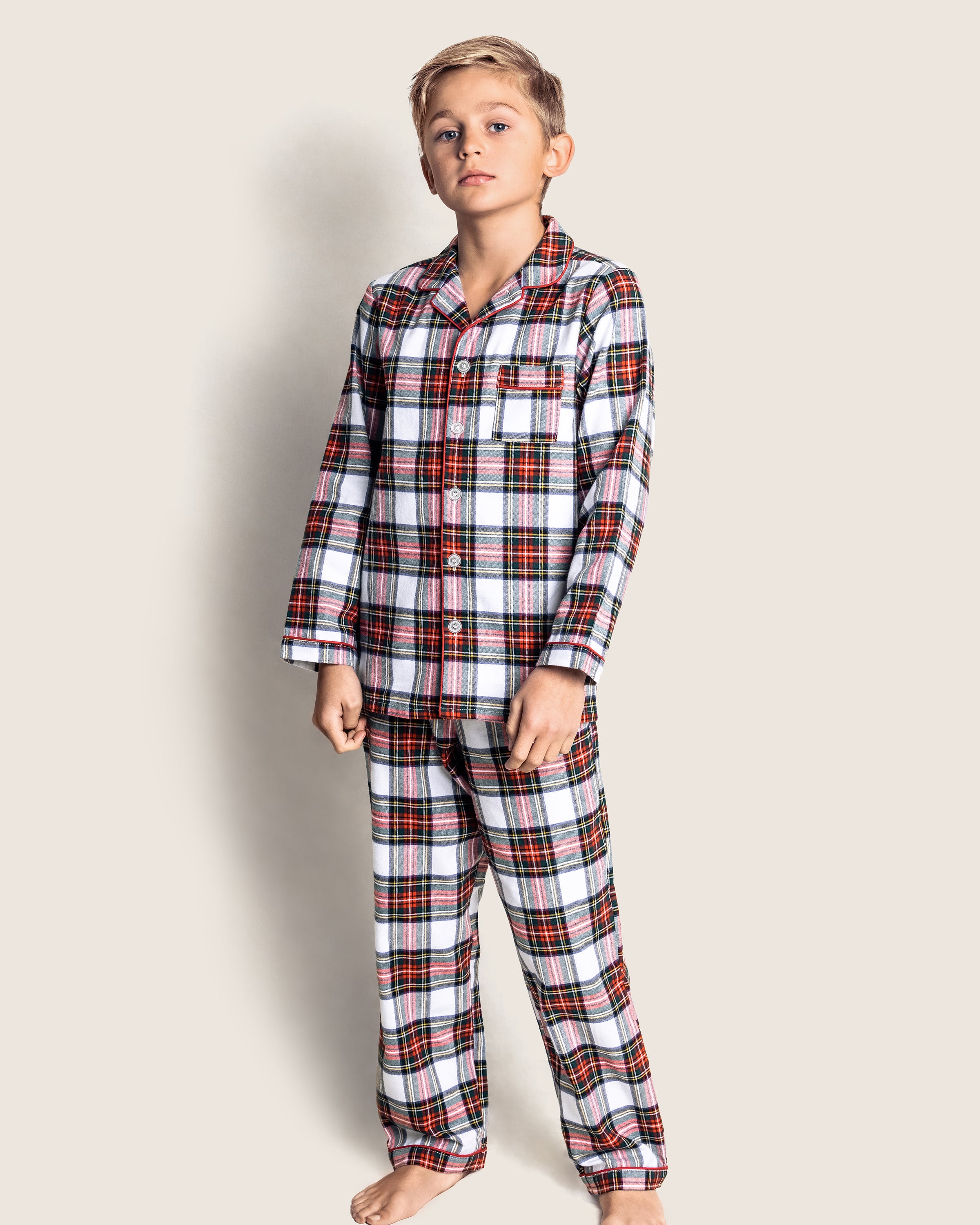 A boy, barefoot against an off-white backdrop, models the Kids Brushed Cotton Pajama Set in Balmoral Tartan by Petite Plume, featuring a plaid pattern with red, black, and green stripes on white. Crafted from soft yarn-dyed cotton for timeless comfort.