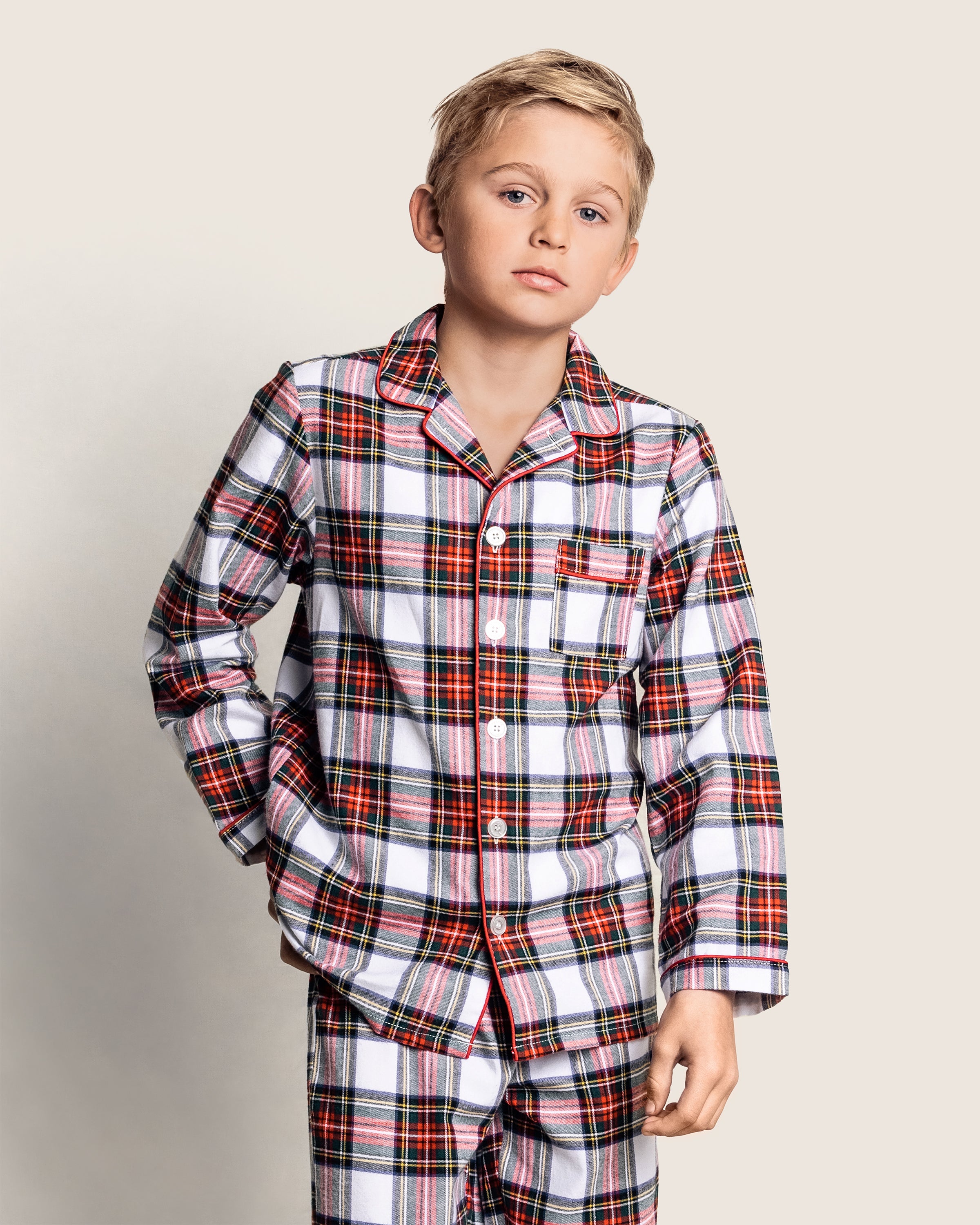 A boy in a Petite Plume Kids Brushed Cotton Pajama Set in Balmoral Tartan, showcasing a white, red, and black checkered pattern, stands with one hand in his pocket against a light background.