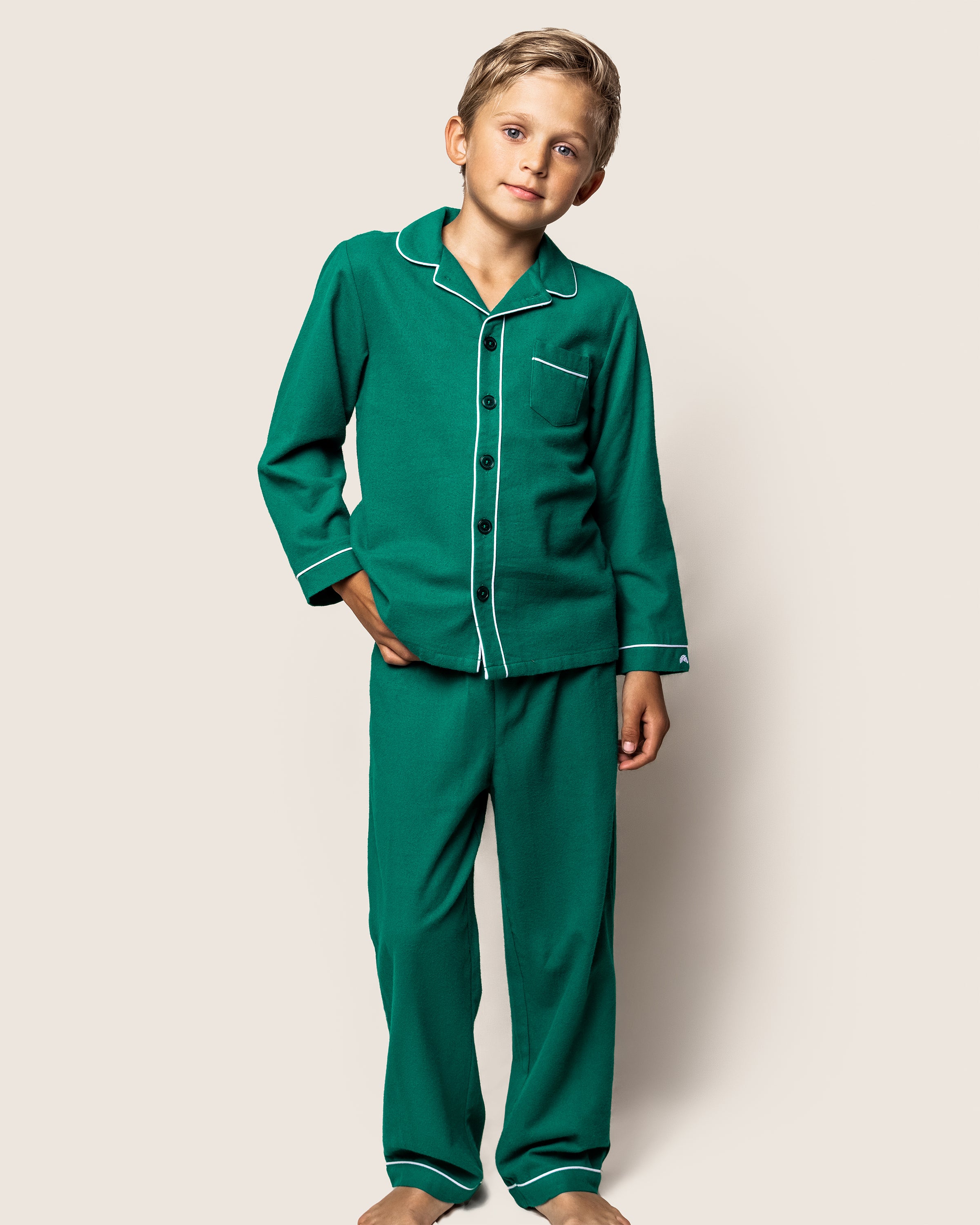 A young boy stands against an off-white background, wearing the Petite Plume Kids Flannel Pajama Set in Forest Green with white piping and black buttons. His right hand is in his pocket, and he has short, light brown hair.