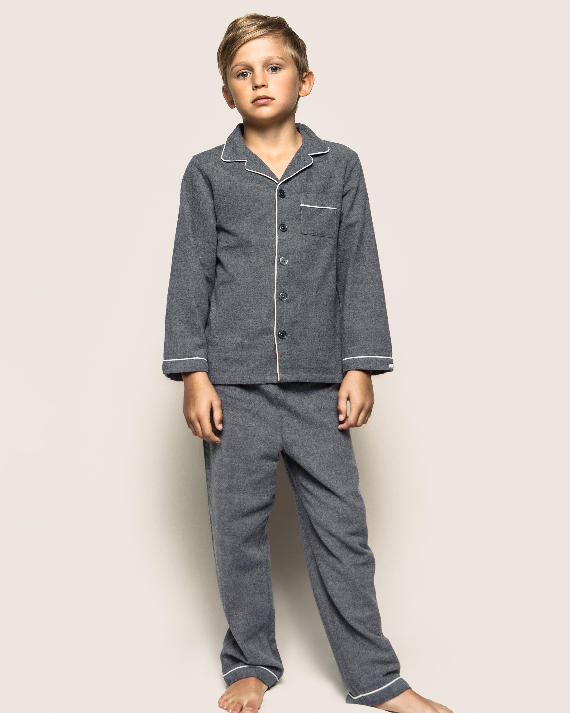 A young boy stands barefoot against a plain background, wearing Petite Plumes Kids Flannel Pajama Set in Grey, designed with white piping and flame-retardant fabric for safety and comfort. His expression is neutral with arms relaxed at his sides.