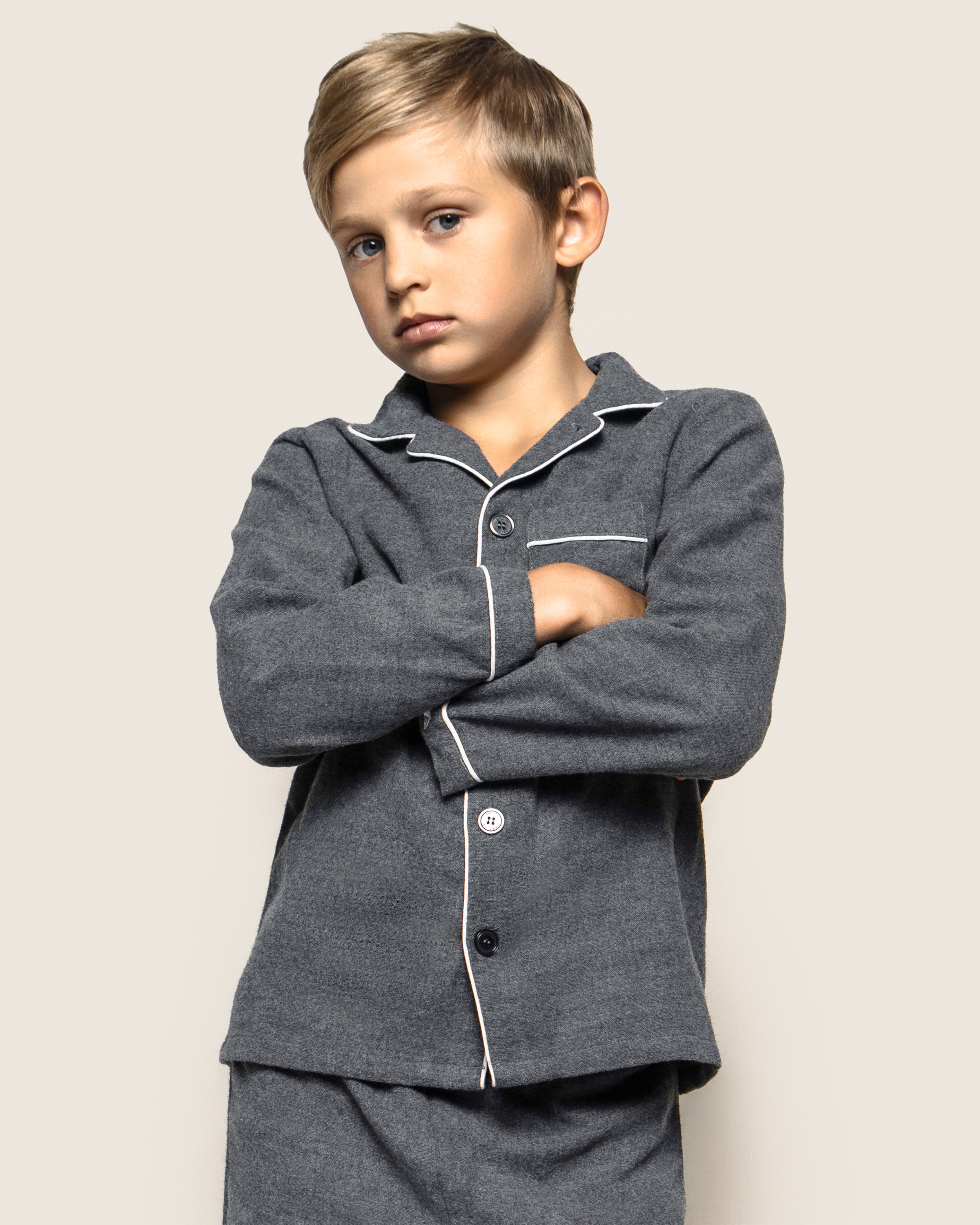 A young boy with short blond hair stands with arms crossed, wearing Petite Plumes Kids Flannel Pajama Set in Grey, featuring white piping. He gazes slightly to the side with a serious expression against a plain background. The sleepwear is crafted from chemical-free materials.