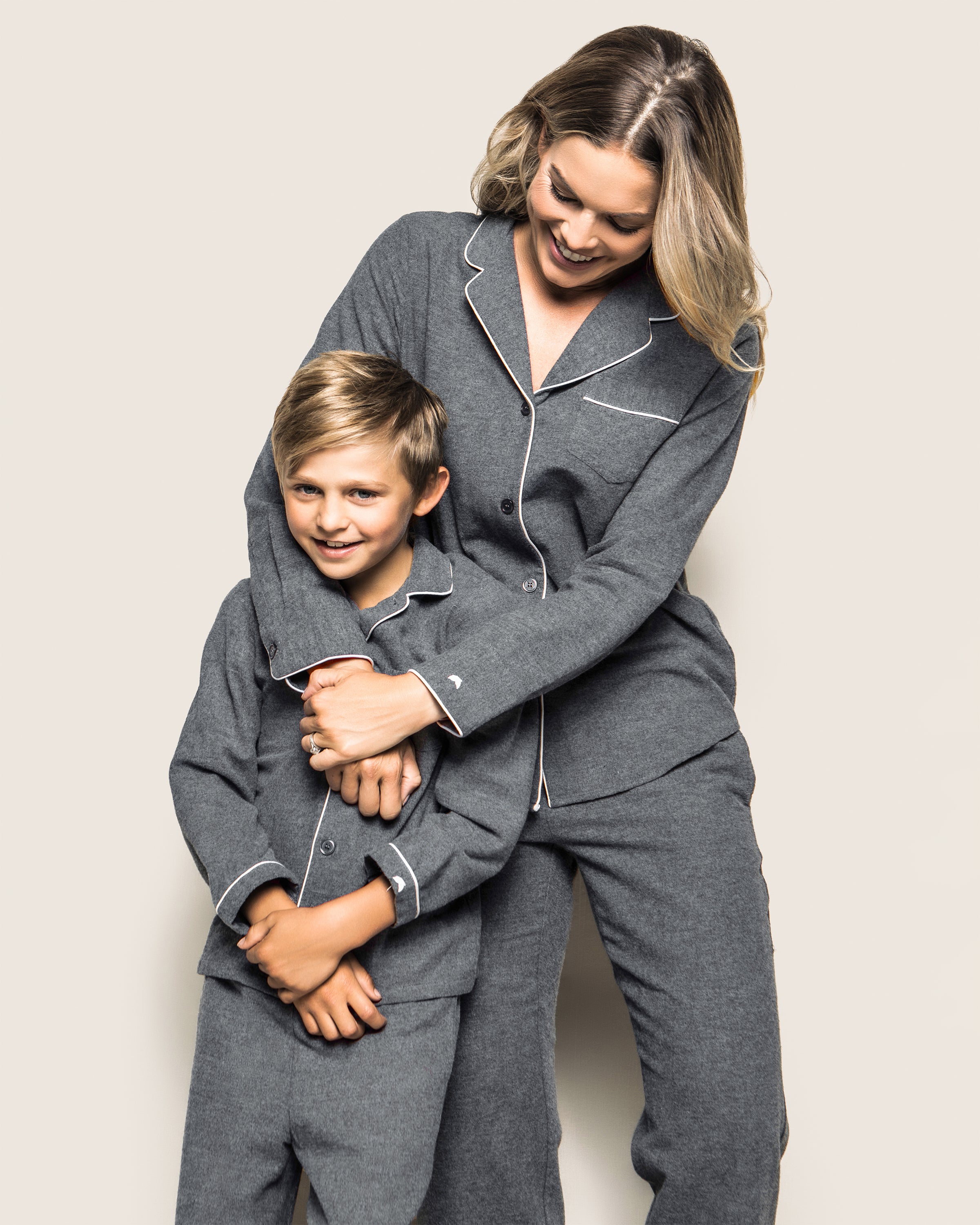 A woman and child in matching flannel PJs from Petite Plume smile together. The woman wraps her arms around the boy, who looks delightedly at the camera. They wear Kids Flannel Pajama Set in Grey against a plain off-white backdrop.