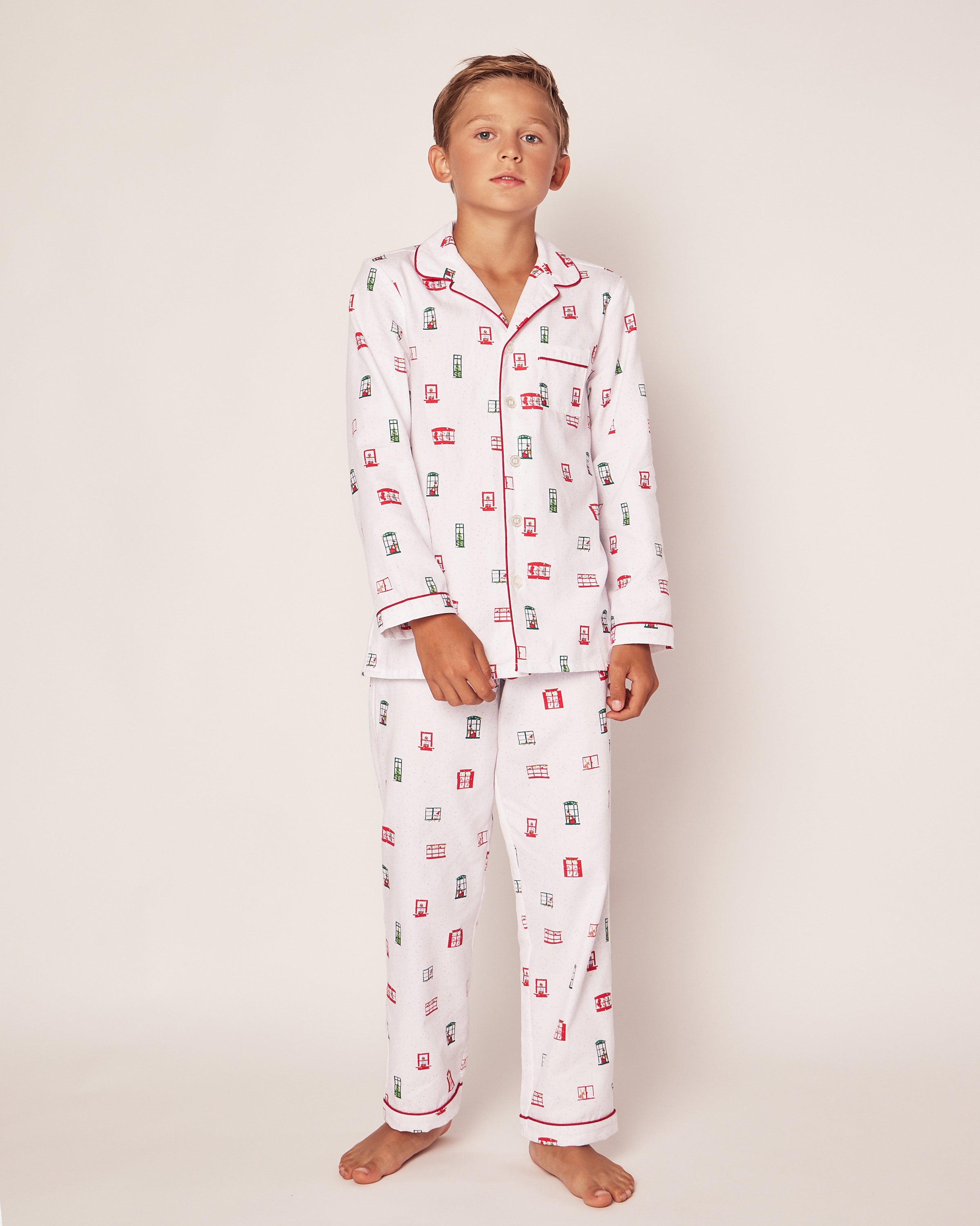 A young boy stands against a plain background in the luxurious Petite Plume Sleepwear Set, featuring a red and green Night Before Christmas print. The pajamas are part of the Maisonette collaboration and include red trim along the collar and sleeves. Hes barefoot, looking relaxed at the camera.