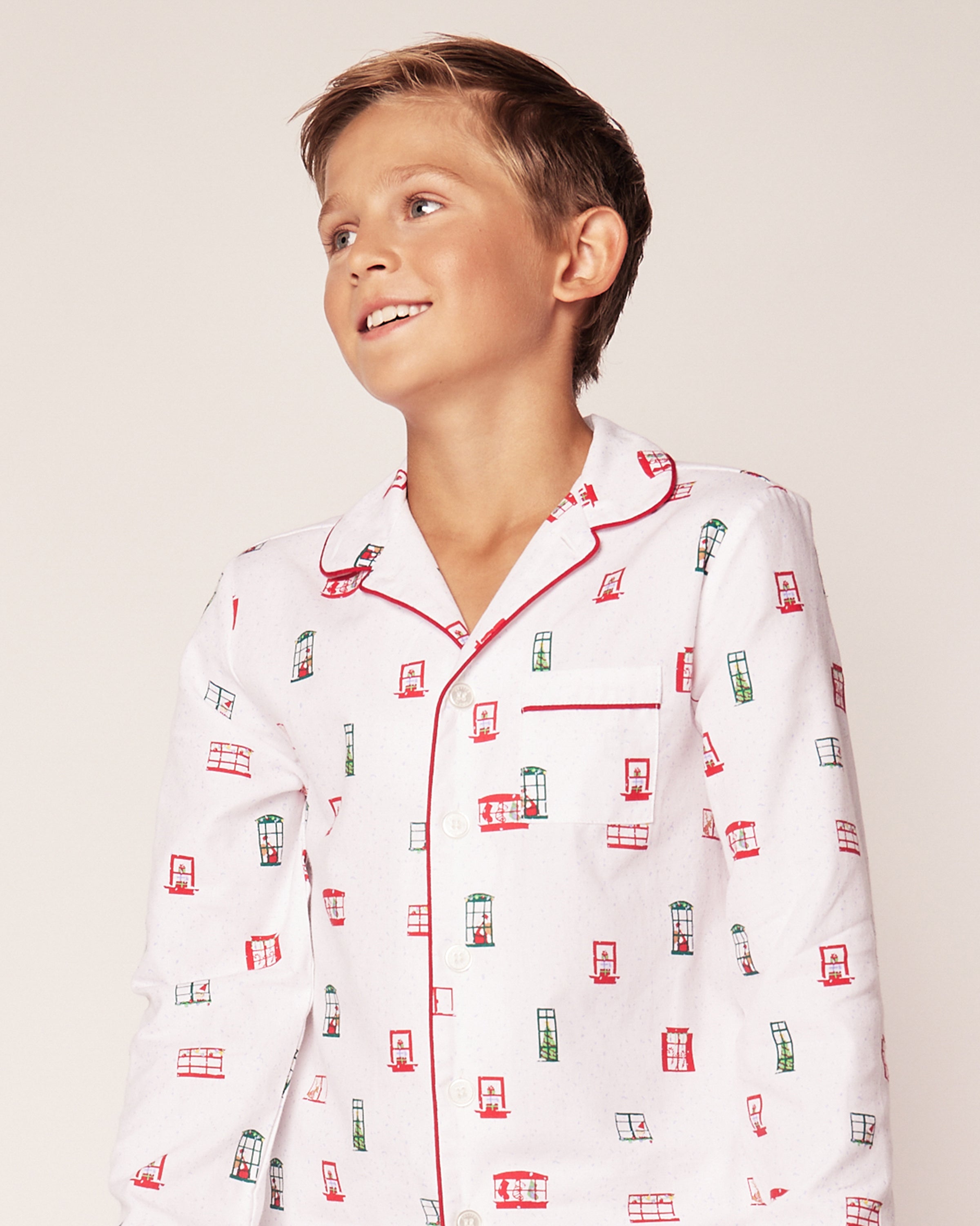 A young boy smiles while wearing the Kids Maisonette x Petite Plume Night Before Pajama Set by Petite Plume, a classic twill pajama set with white fabric adorned in a whimsical Night Before Christmas red trim and small red and green window designs.