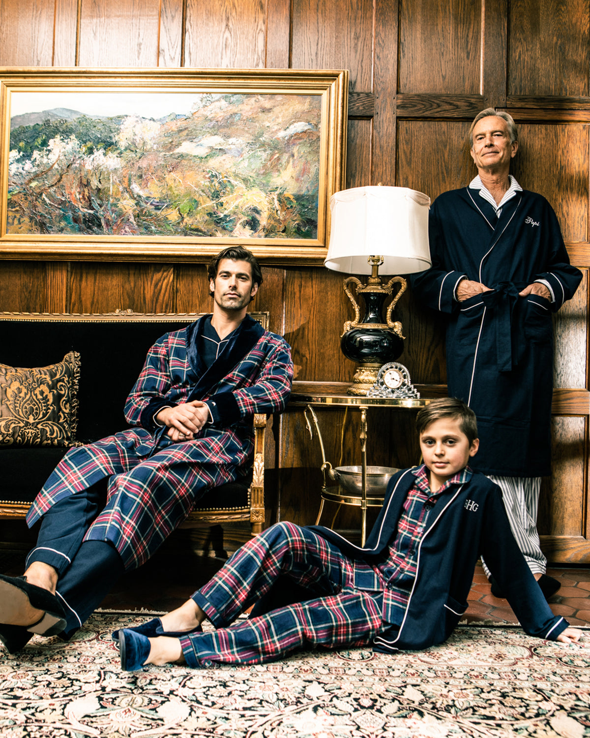 Three people in matching Kid’s Brushed Twill Pajama Sets in Windsor Tartan and navy robes relax in a wood-paneled room with an ornate rug, a plush sofa, a lamp on a side table, and a framed landscape painting on the wall behind them.