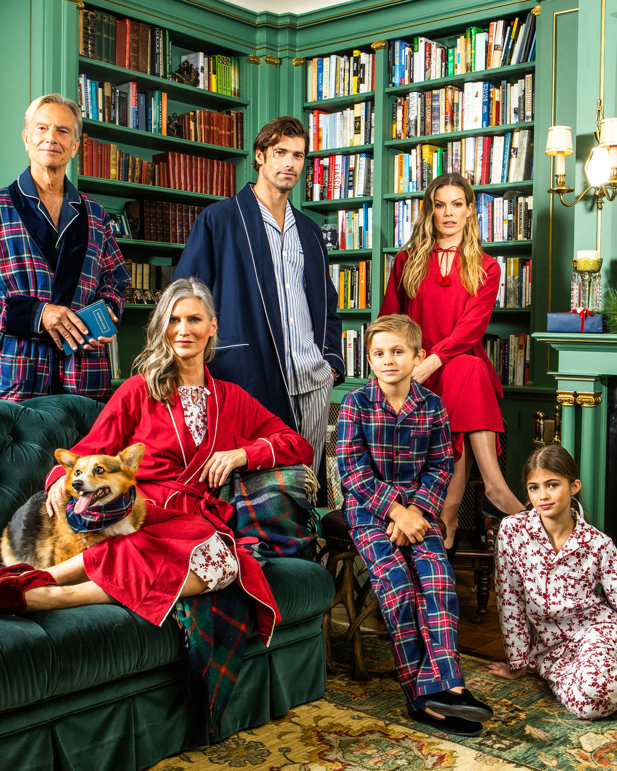 Six people in festive pajamas and robes pose together in a cozy, book-filled library. A dog sits on the green sofa next to a woman with gray hair. One child wears the Kid’s Brushed Twill Pajama Set in Windsor Tartan, adding to the group’s relaxed, holiday feel.
