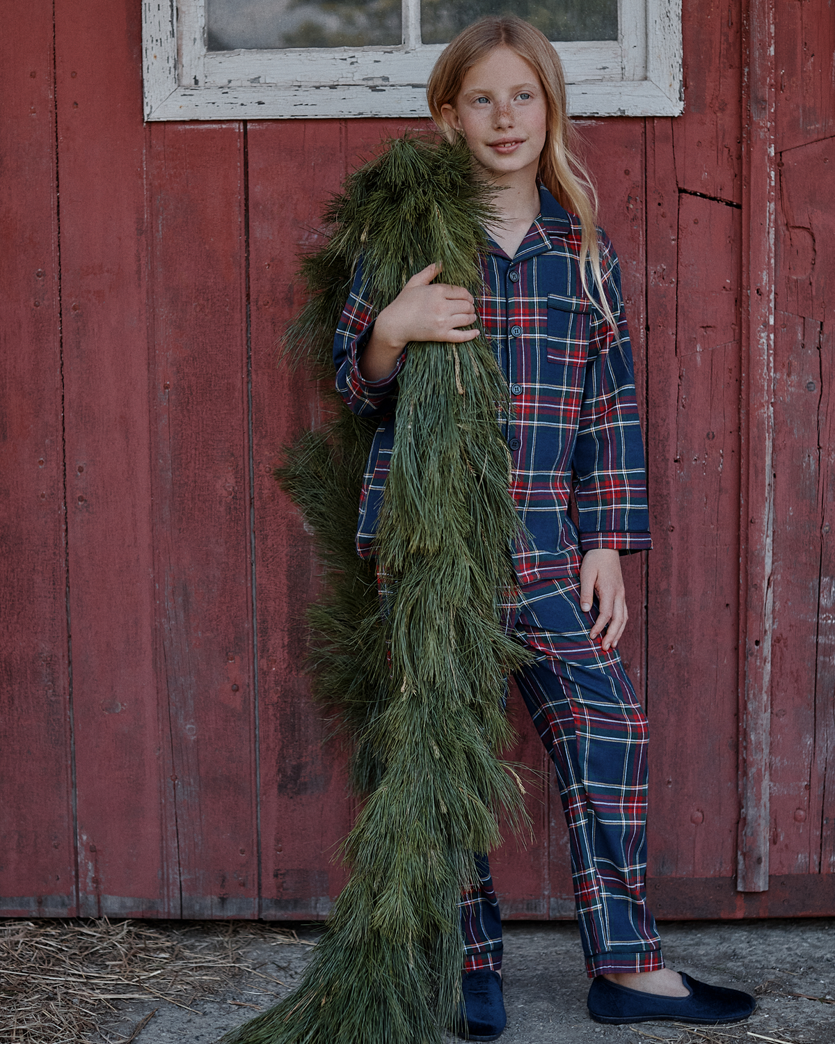 A young girl wearing the Kid’s Brushed Twill Pajama Set in Windsor Tartan stands by a red wooden wall, holding a long, green pine garland draped over her shoulder.