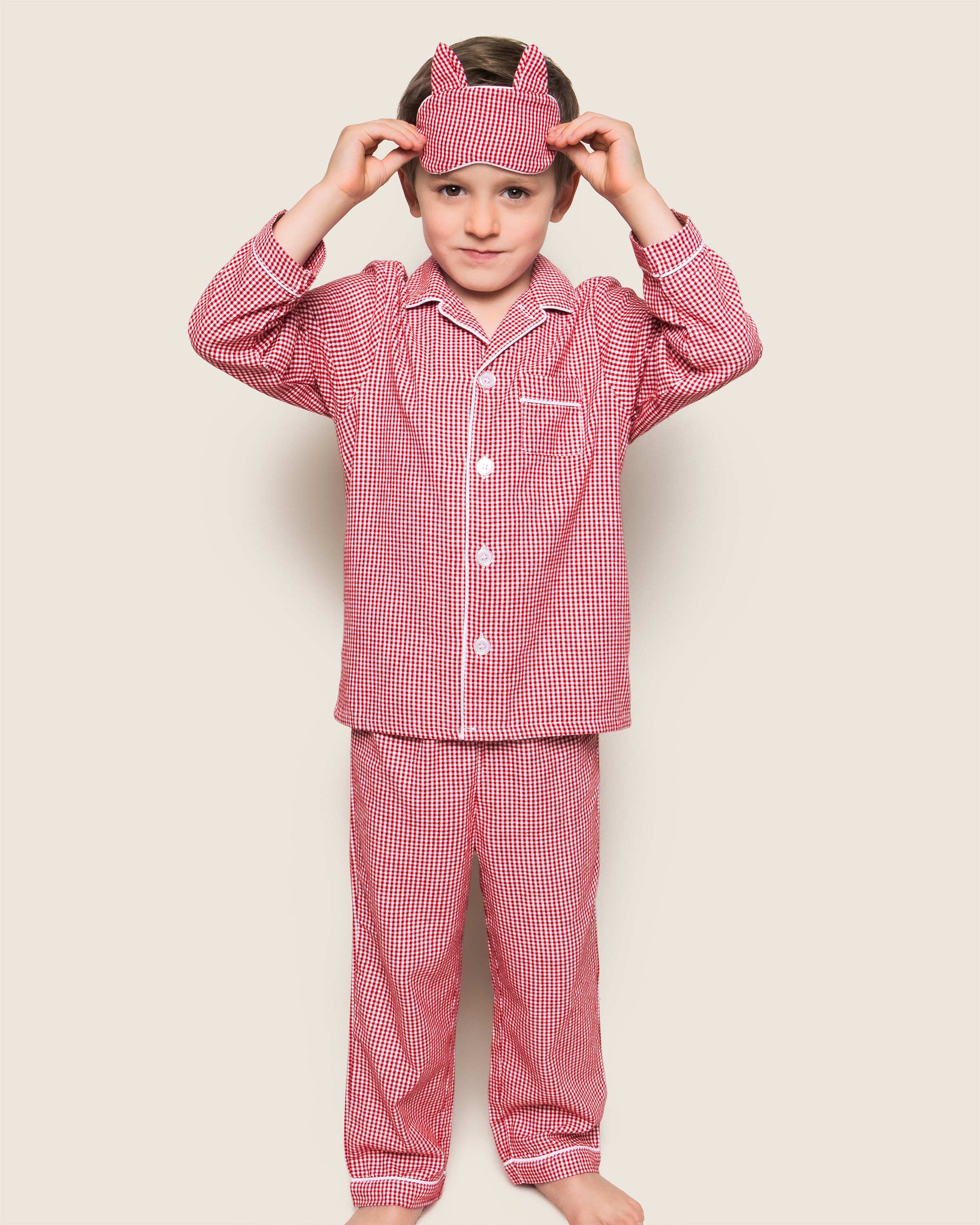 A child, in Petite Plumes Red Mini Gingham Kids Flannel Pajama Set, stands barefoot against a neutral background adjusting a matching sleep mask.