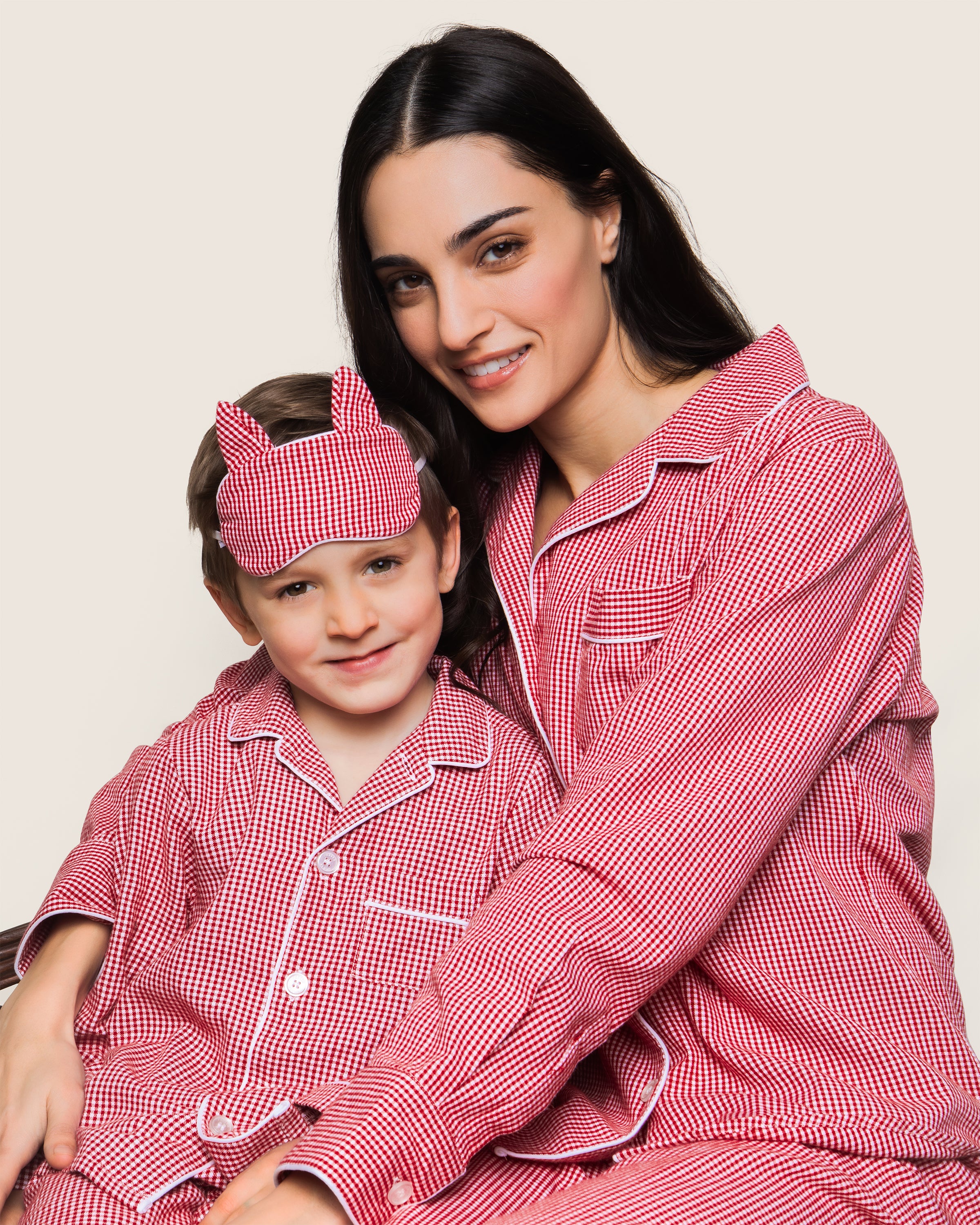 A woman and child happily sit together in Petite Plumes matching flannel pajamas, with the child sporting a Red Mini Gingham sleep mask. They embody the warmth of a classic set against a plain backdrop.
