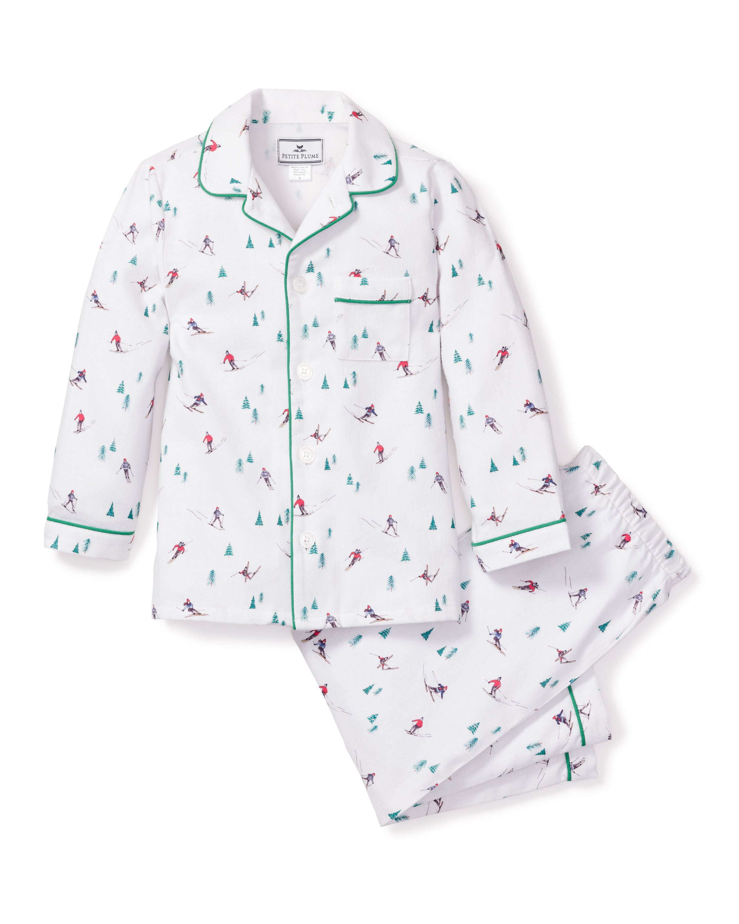 Petite Plumes Kids Flannel Pajama Set in Après Ski features a white skiing pattern with small figures and trees. The button-down top has green piping on the collar and cuffs, while the matching pants have an elastic waistband. Made from flame-retardant fabric for added safety and comfort.