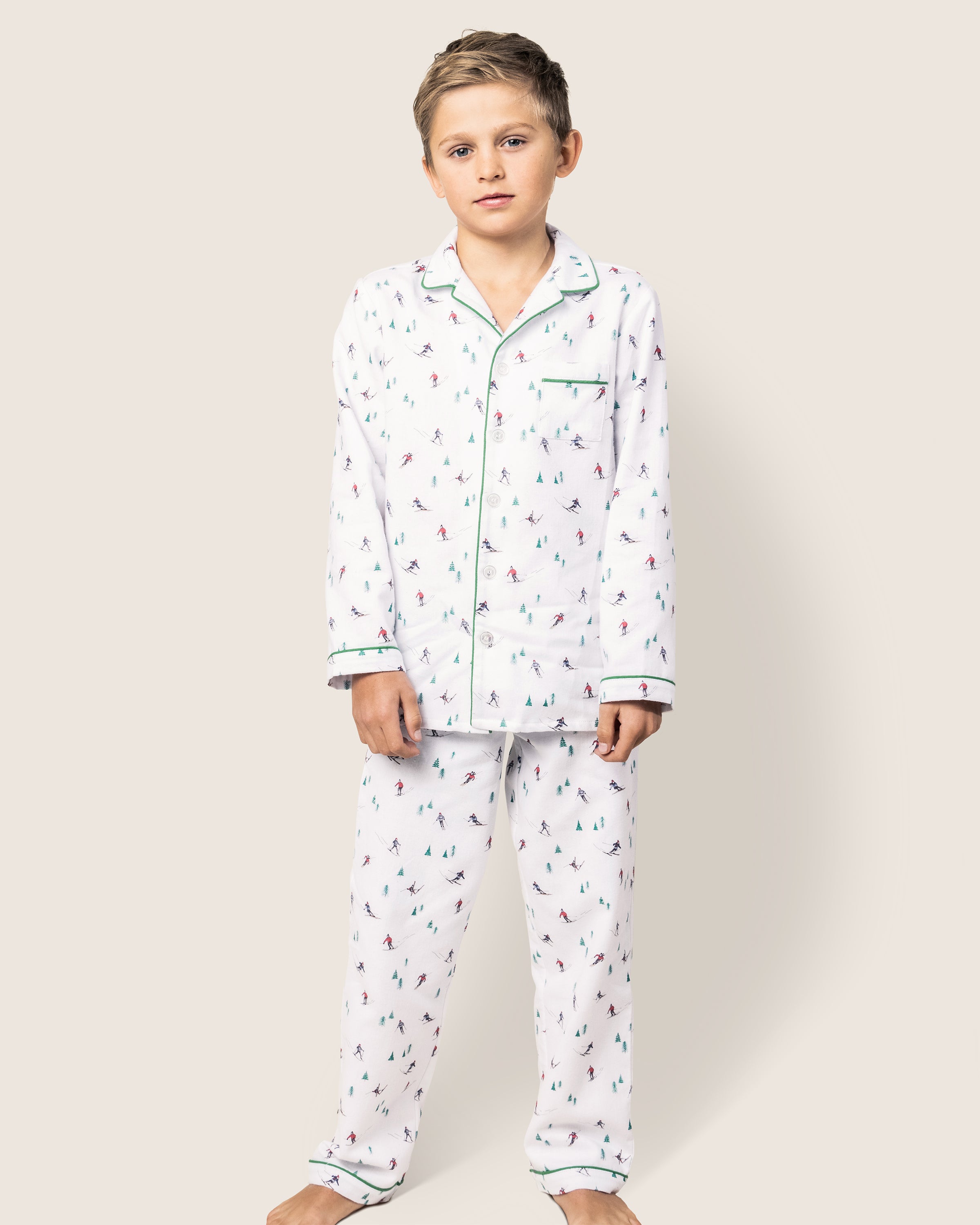 A young boy stands barefoot against a plain background, wearing Petite Plumes cozy Kids Flannel Pajama Set in Après Ski. These flame-retardant pajamas feature colorful designs, a classic collar, and front buttons for a charming look.
