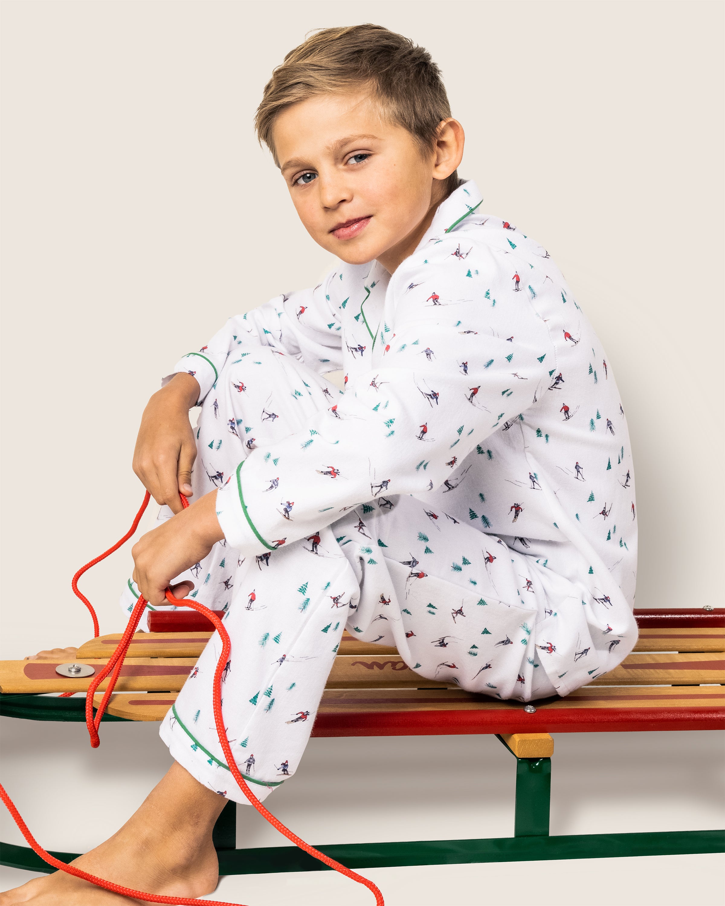 A young boy in Petite Plumes Kids Flannel Pajama Set in Après Ski sits on a red and green sled, clutching its red rope, looking relaxed against a neutral background.