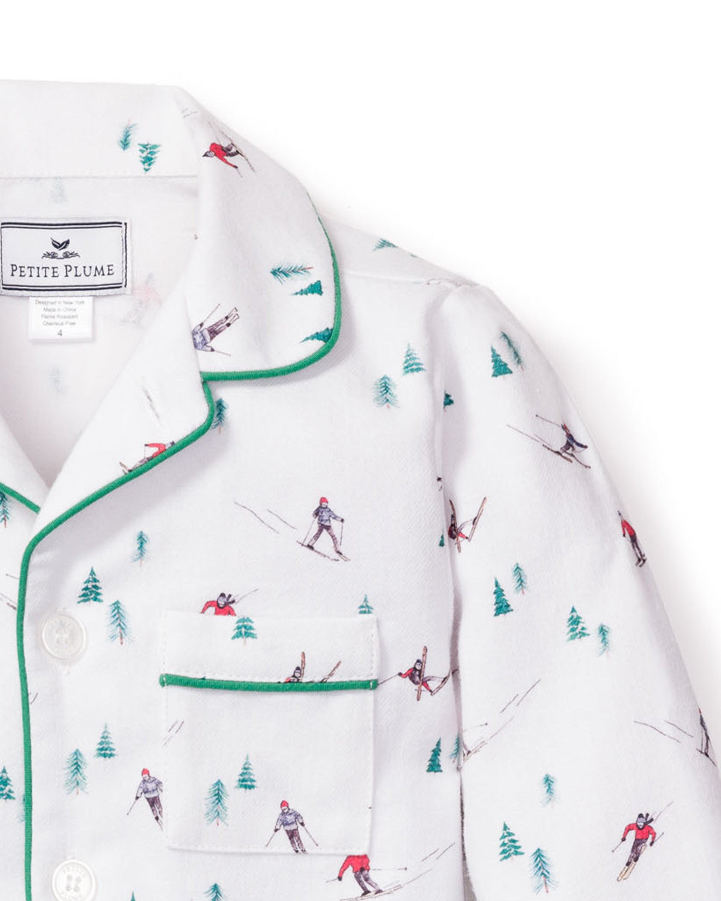 Petite Plumes Kids Flannel Pajama Set in Après Ski features white flannel adorned with skiers and green trees, green piped trim, and a chest pocket. The collar label assures quality comfort with flame-retardant fabric.