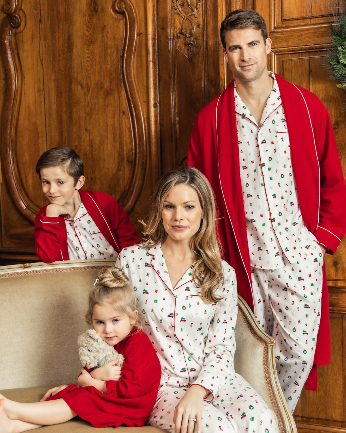 A family of four in festive Christmas pajamas poses together indoors. The father and son stand at the back in soft cotton twill pajamas with red robes, while the mother sits on the couch wearing Petite Plumes Womens Twill Pajama Set in Winter Nostalgia next to her daughter in a red nightgown.