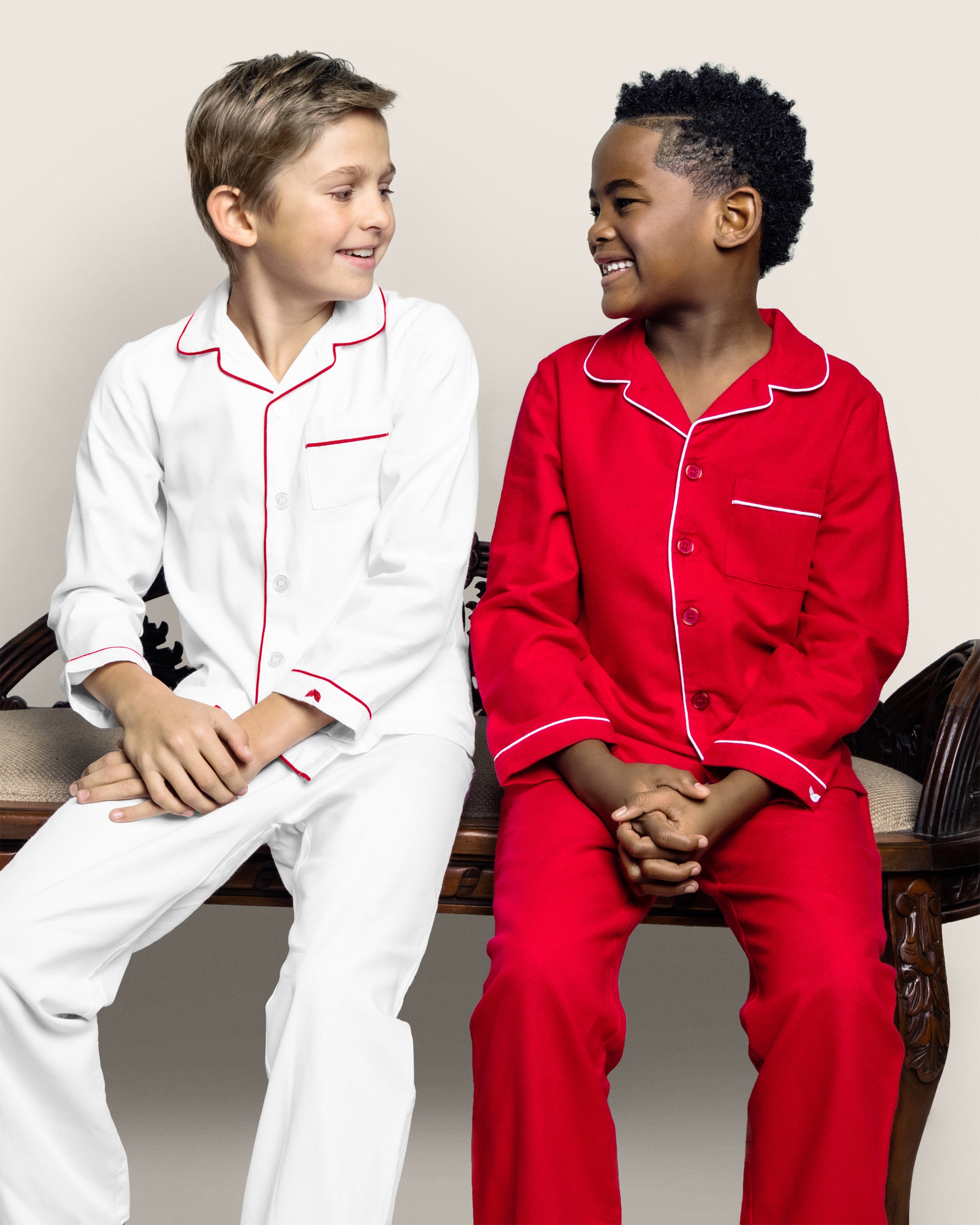 Two children sit on a bench, smiling in classic pajama sets. One wears Petite Plume’s Kids Twill Pajamas in White with Red Piping, while the other sports red pajamas with white trim. The cozy scene unfolds against a plain light gray background.