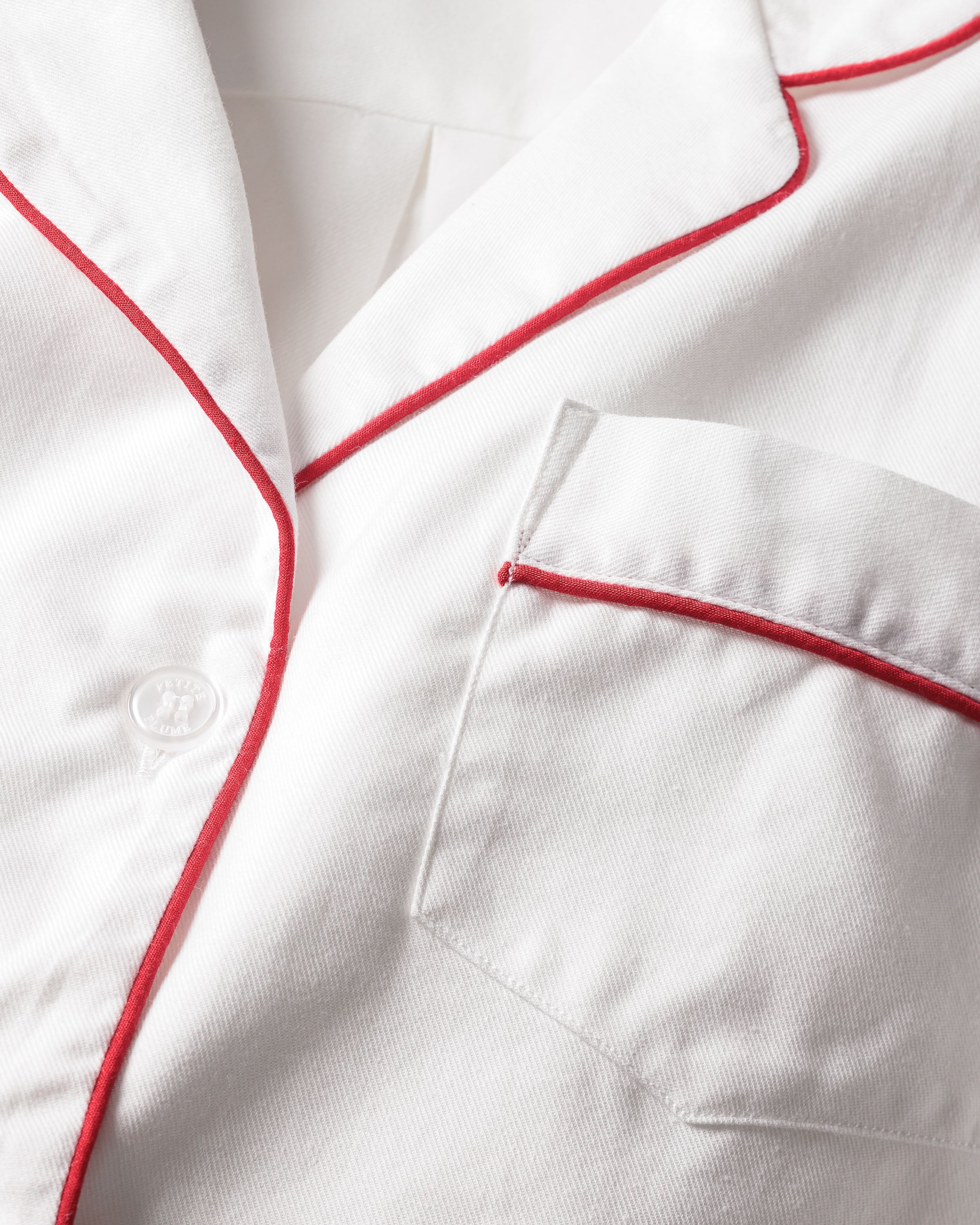 A close-up of Petite Plumes classic Kids Twill Pajamas in white fabric showcases a collar with red piping, a single button, and a right-side pocket outlined with red piping, adding subtle contrast to the sleepwear.
