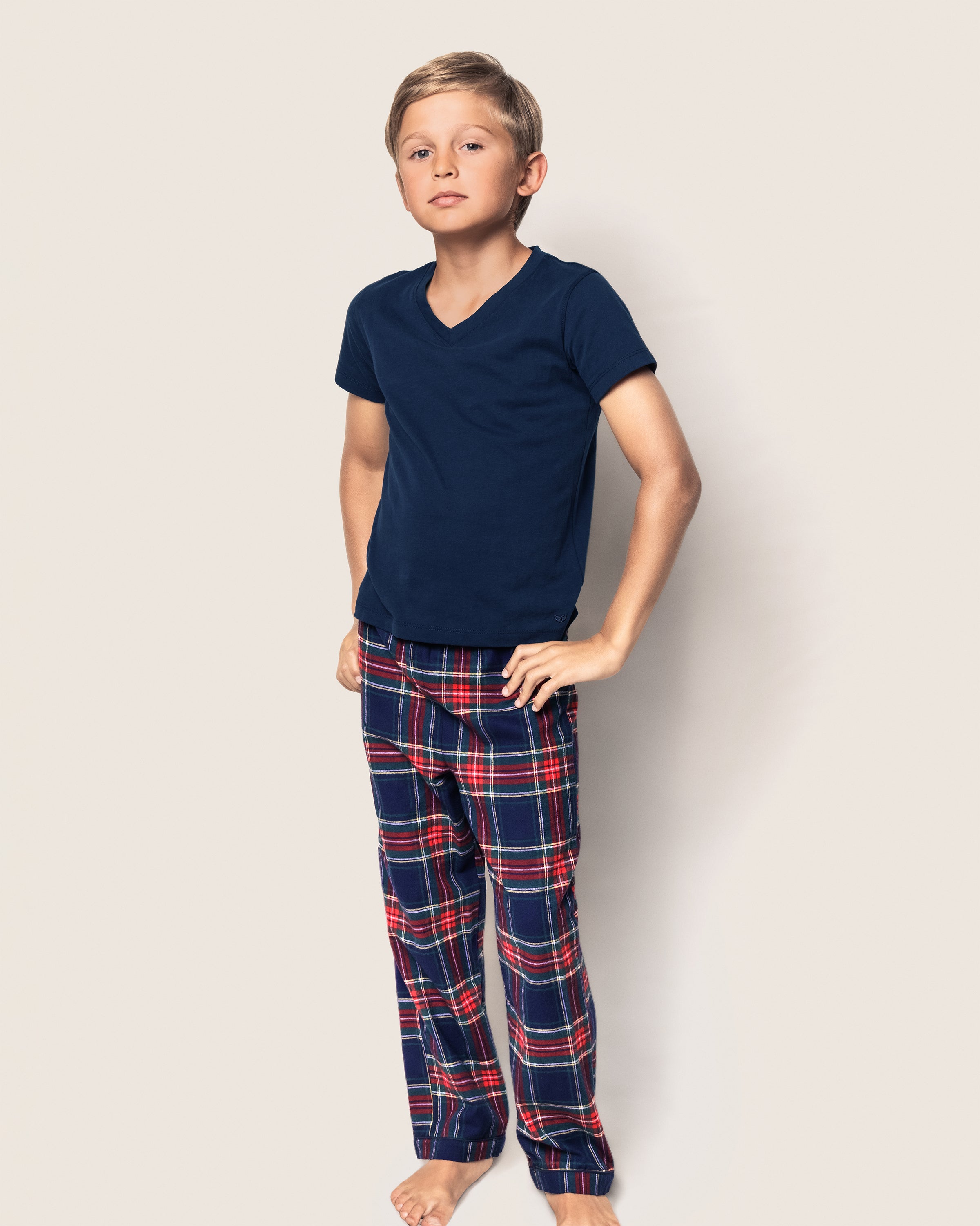 A boy with short hair confidently stands against a plain background, wearing a navy blue T-shirt and Petite Plumes Kids Brushed Cotton Pajama Pant in Windsor Tartan. His hands rest on his hips while he looks at the camera.