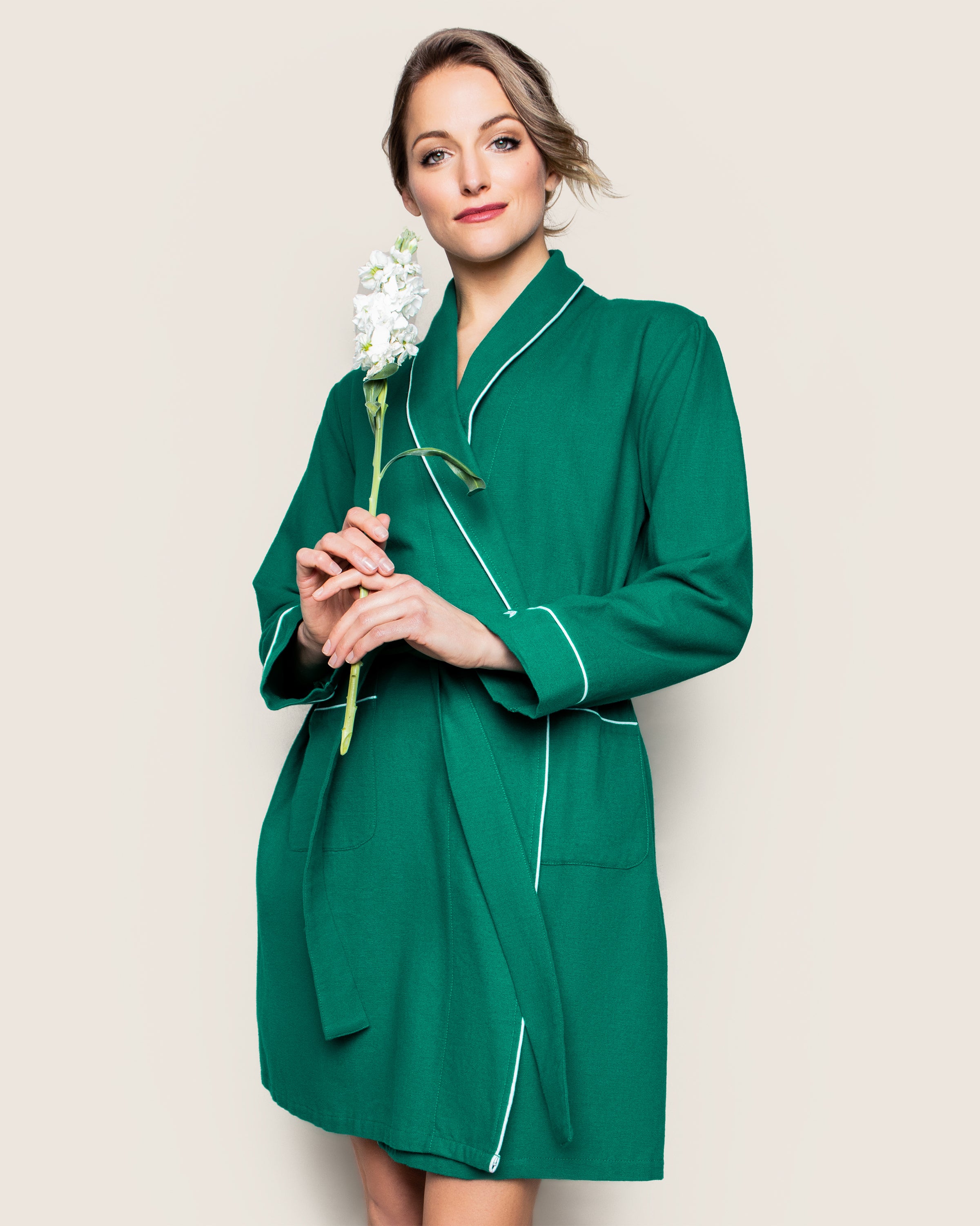 A person wearing a Petite Plume Womens Flannel Robe in Forest Green with white trim holds a white flower while standing against a plain background. They have neatly styled short hair and are looking directly at the camera.