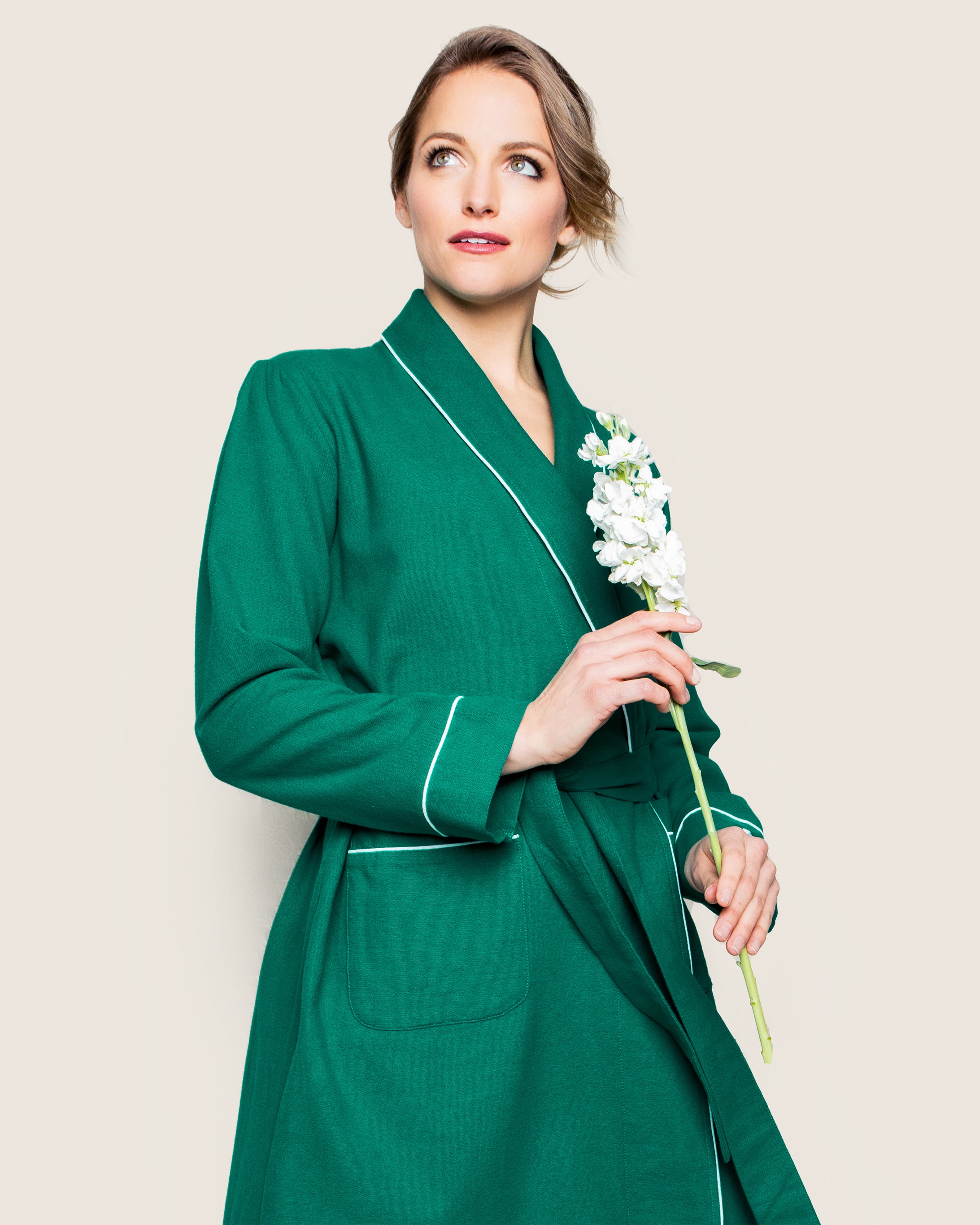 A person in a Womens Flannel Robe in Forest Green by Petite Plume gently holds a white flower. Their light brown hair is elegantly styled in an updo as they gaze to the side against a plain beige background.