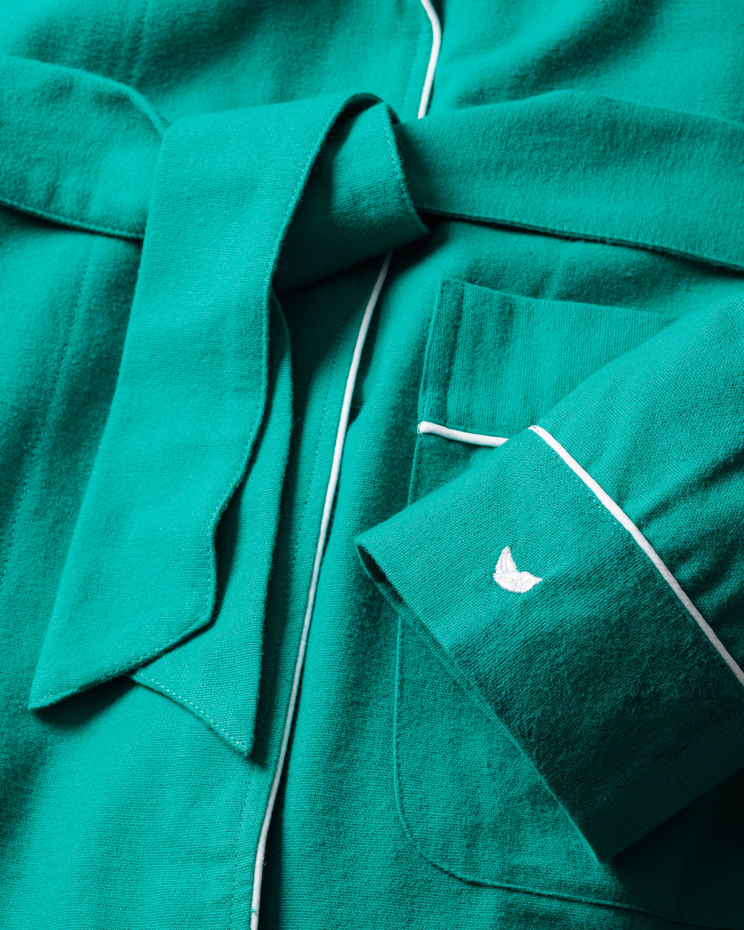Close-up of a forest green flannel robe by Petite Plume, featuring a wide belt tied at the front. Crafted from soft cotton twill, it showcases white piping along the edges and a small white embroidered logo on the pocket and sleeve cuff.