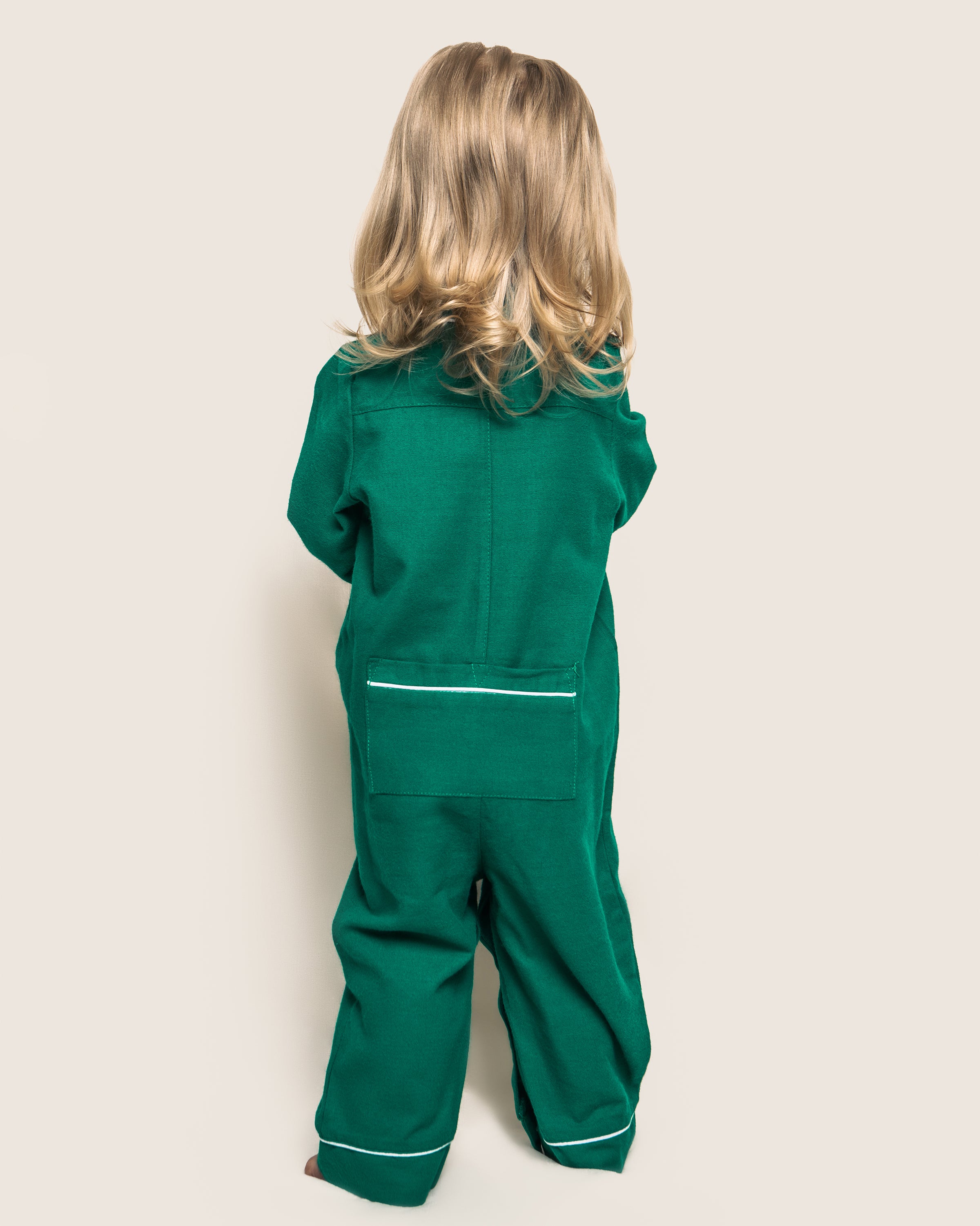 A child with long blonde hair faces away, wearing Petite Plumes Babys Flannel Romper in Forest Green with White Piping. The light cream background subtly contrasts with the cozy sleepwear feel.