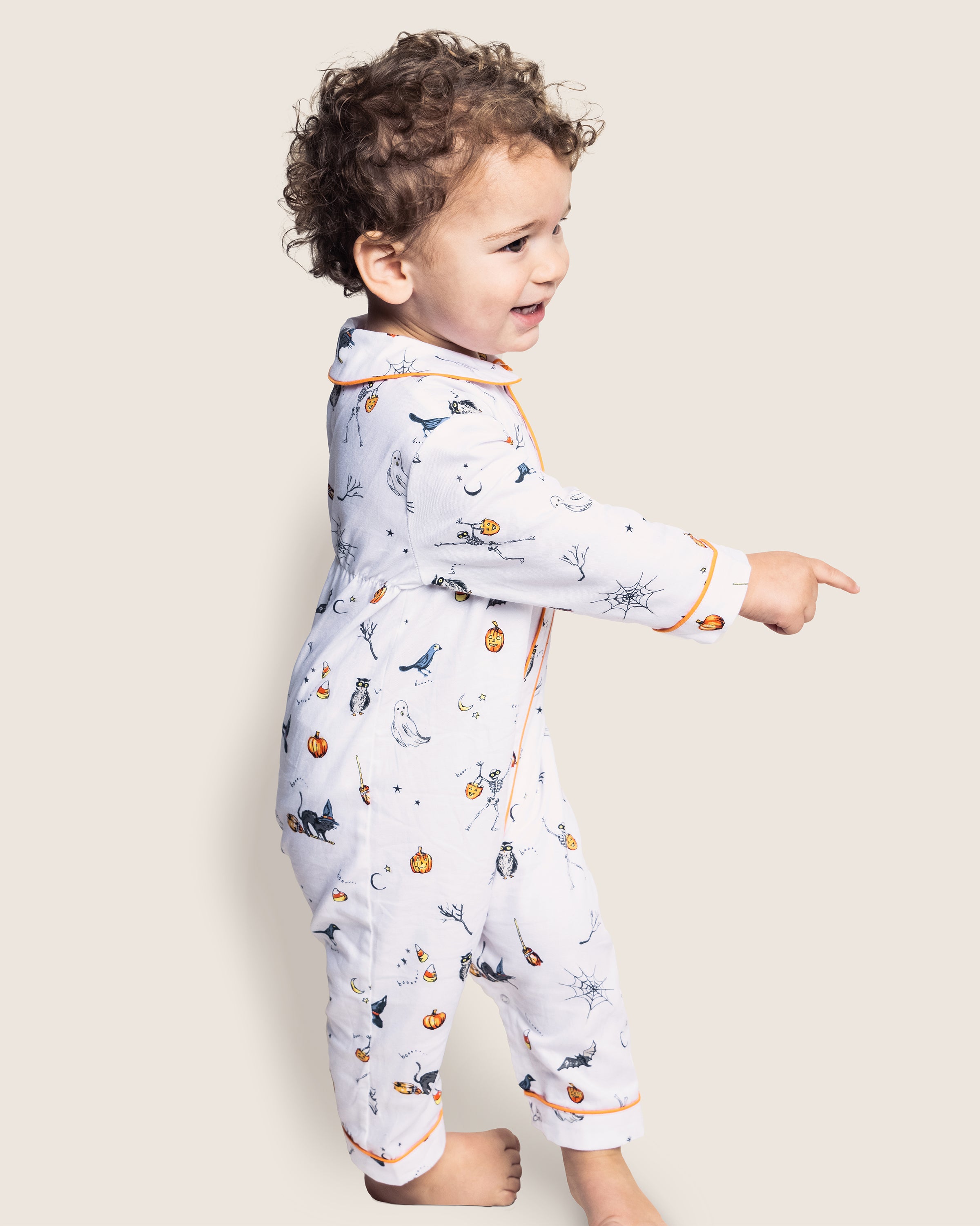 A toddler in Petite Plume’s Babys Twill Cambridge Romper in Trick or Treat print, showcasing playful pumpkins and leaves, stands barefoot on a light background. With curly hair and a joyful expression, the child points to the right, embodying Halloween icons.