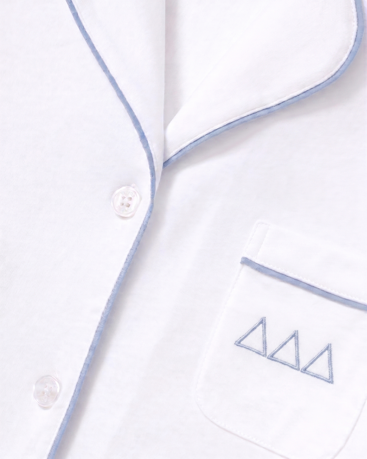 Close-up of the Greek Embroidered Poppy Pima Short Set pajama top in white with periwinkle piping, featuring three buttons and a pocket adorned with three small blue embroidered triangle designs above the trim.