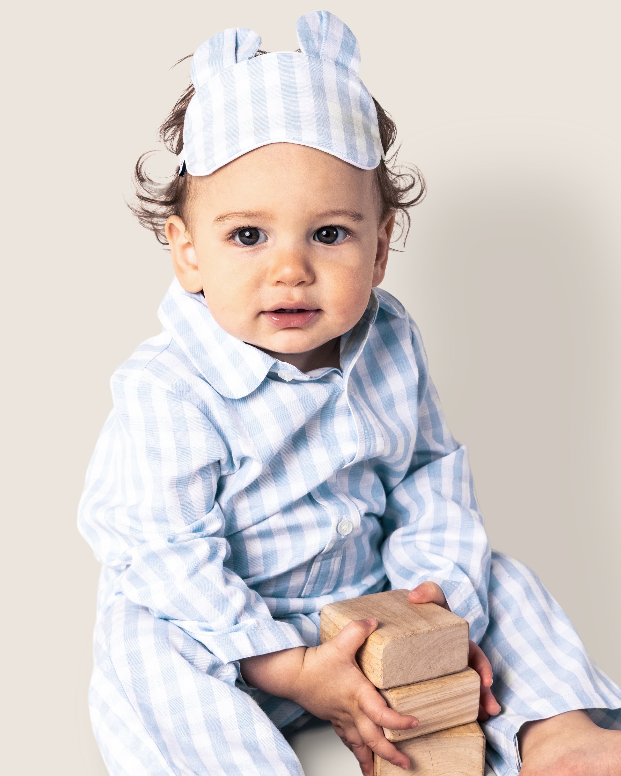 A toddler sits holding wooden blocks in Petite Plumes Babys Twill Romper in Light Blue Gingham, with a bear ear sleep mask on their head. The yarn-dyed cotton ensemble stands out against the soft beige background.