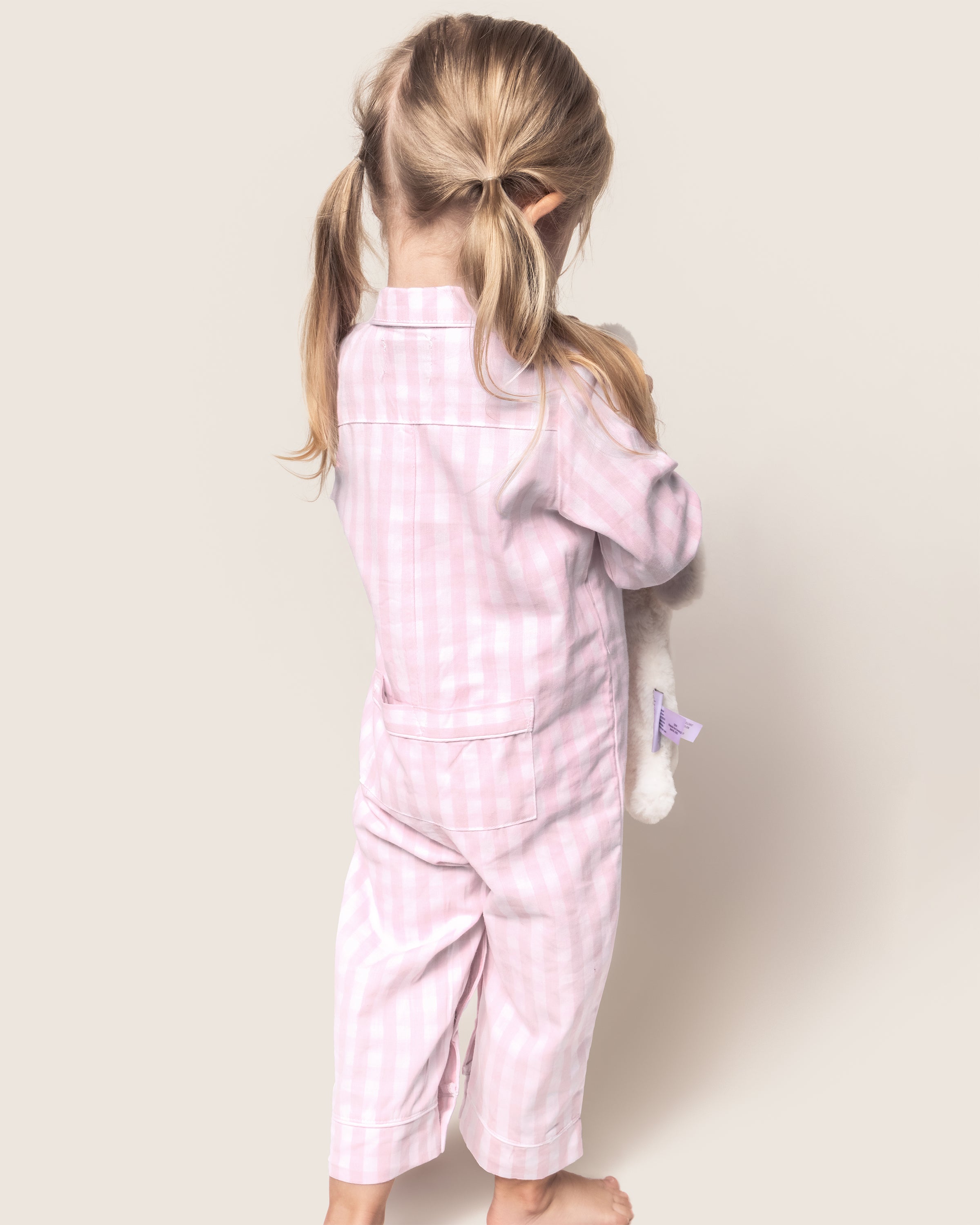 A young girl with pigtails stands barefoot, facing away. She wears Petite Plumes Babys Twill Romper in Pink Gingham made of yarn-dyed cotton for lasting color. She hugs a stuffed animal against a plain background.