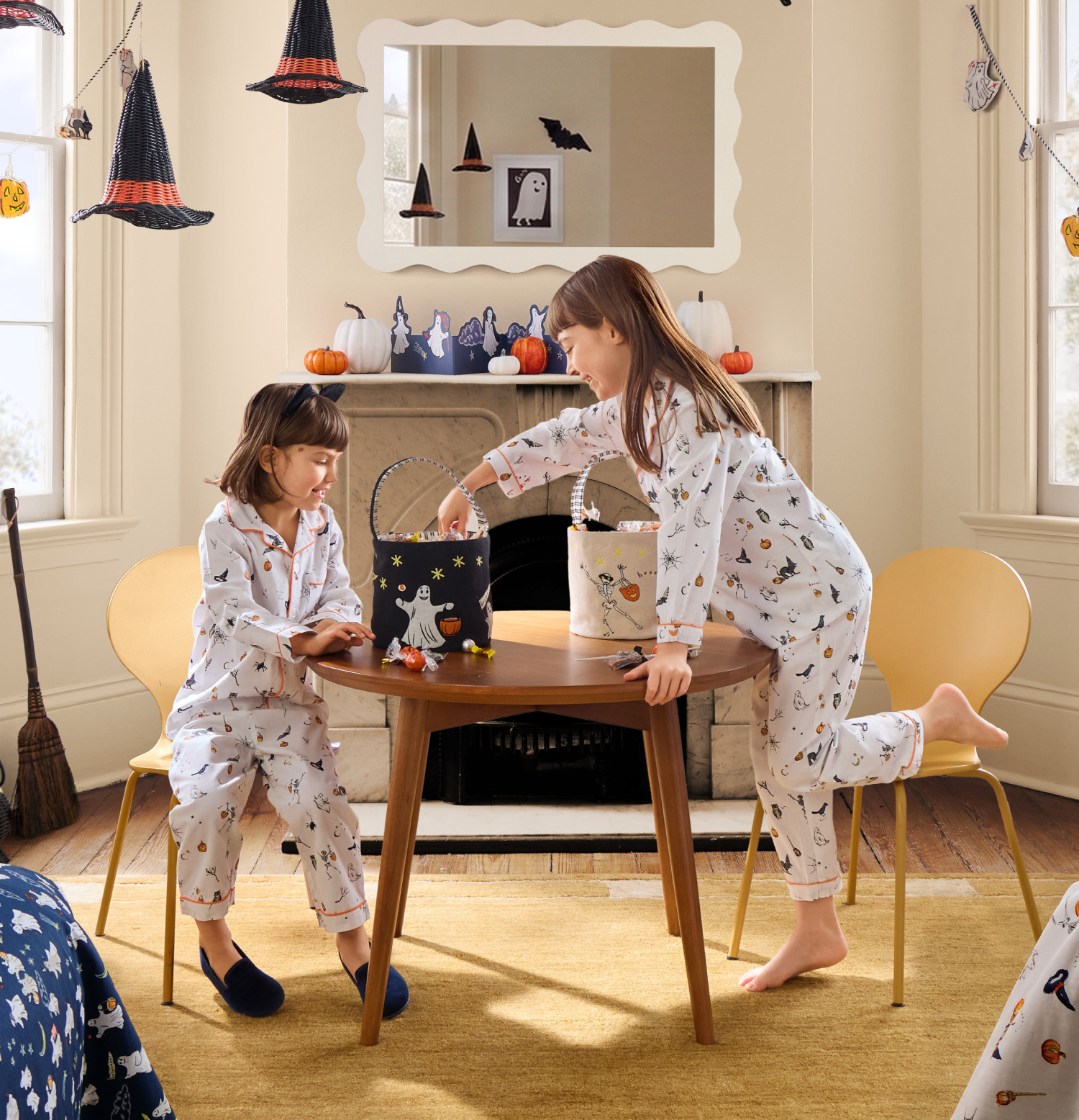 Two children in Halloween pajamas sit and stand at a small table, reaching into treat buckets. The room is decorated with pumpkins, witch hats, and paper bats, giving a festive Halloween atmosphere.