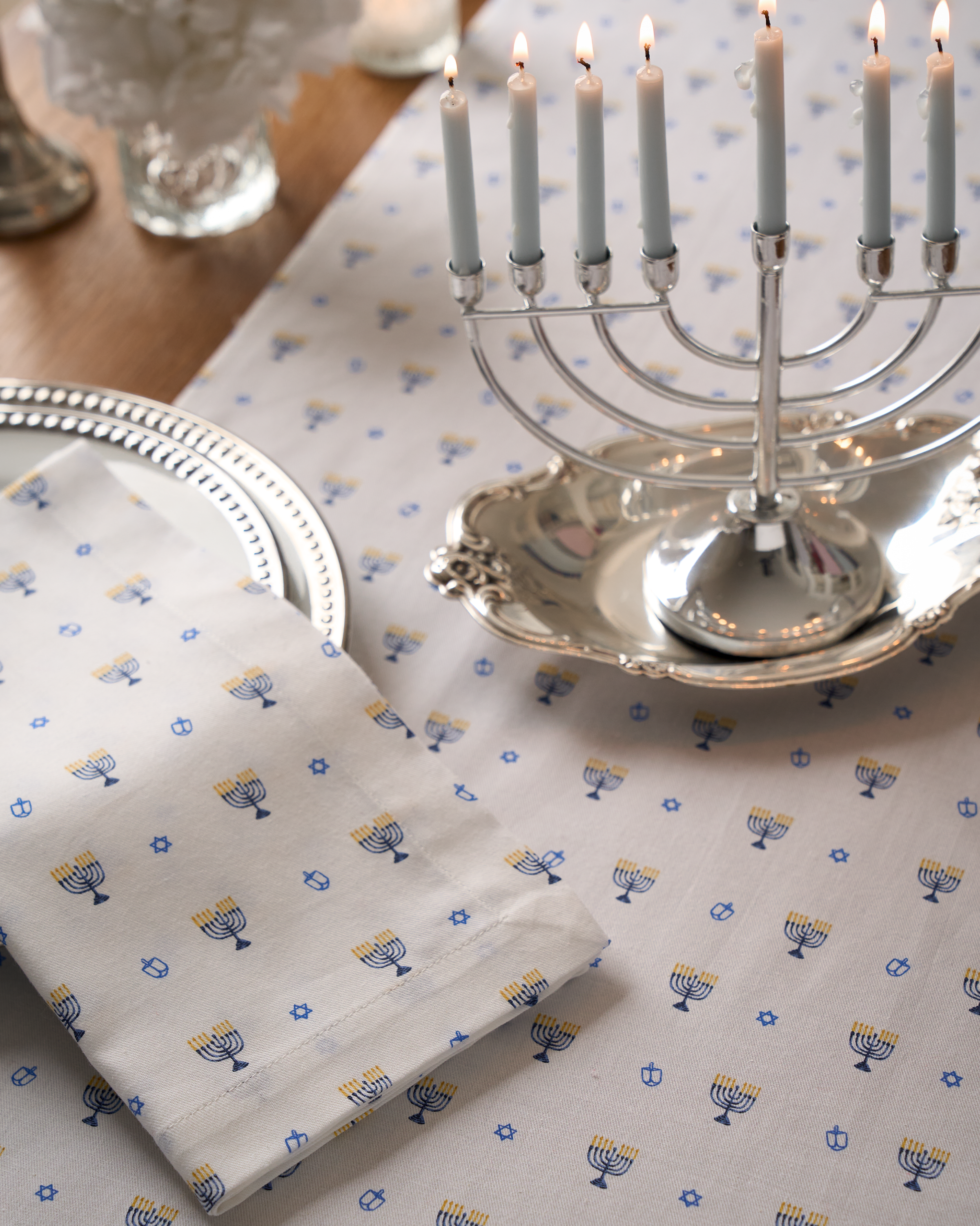 A silver menorah with lit candles sits on a festive tablescape featuring the Signature Twill Table Runner in Happy Hanukkah by Petite Plume. The 100% cotton white tablecloth is adorned with blue and yellow menorahs, stars, and dreidels, paired with matching napkins and silver plates for ambiance.