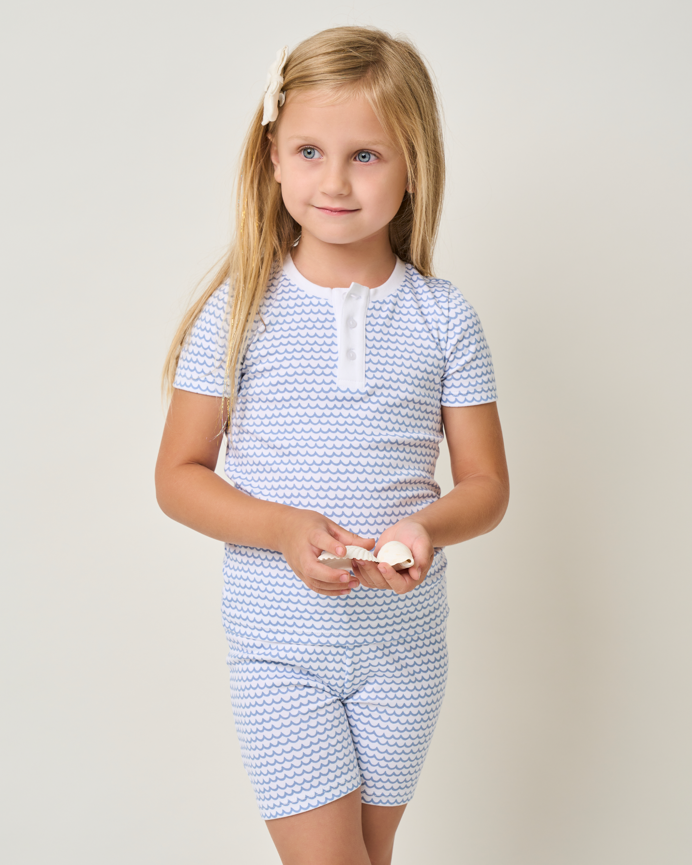 A young girl with long blonde hair stands against a plain background, wearing Petite Plumes Kids Pima Snug Fit Pajama Short Set in La Mer, featuring a small wave pattern. She holds seashells and has a white bow in her hair.