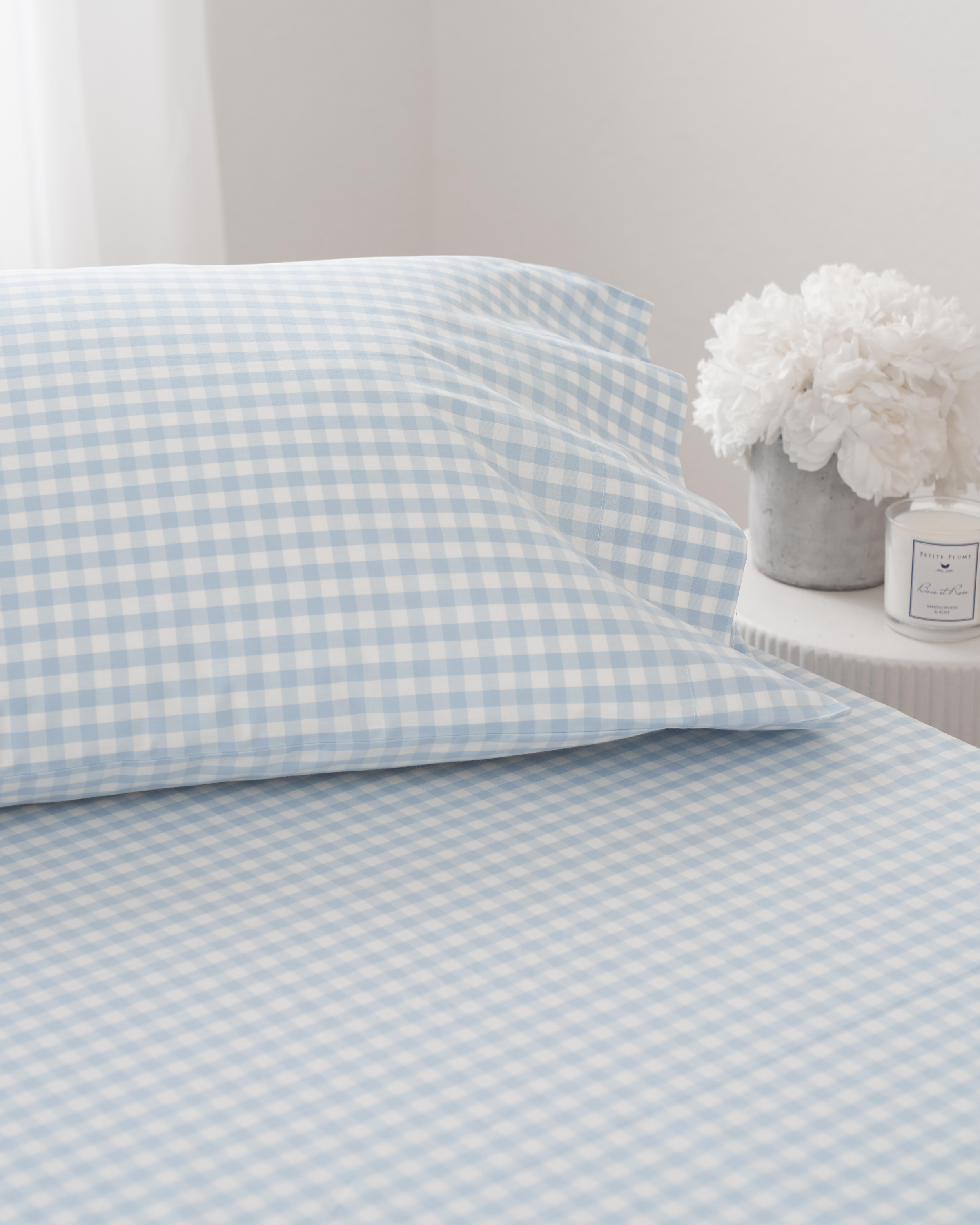 A neatly made bed features the Luxe Premium Cotton Light Blue Gingham Bed Sheets and a matching pillowcase by Petite Plume. A nearby round table holds white flowers and a scented candle, while soft natural light from the window enhances the elegant ambiance of this exquisite setting.