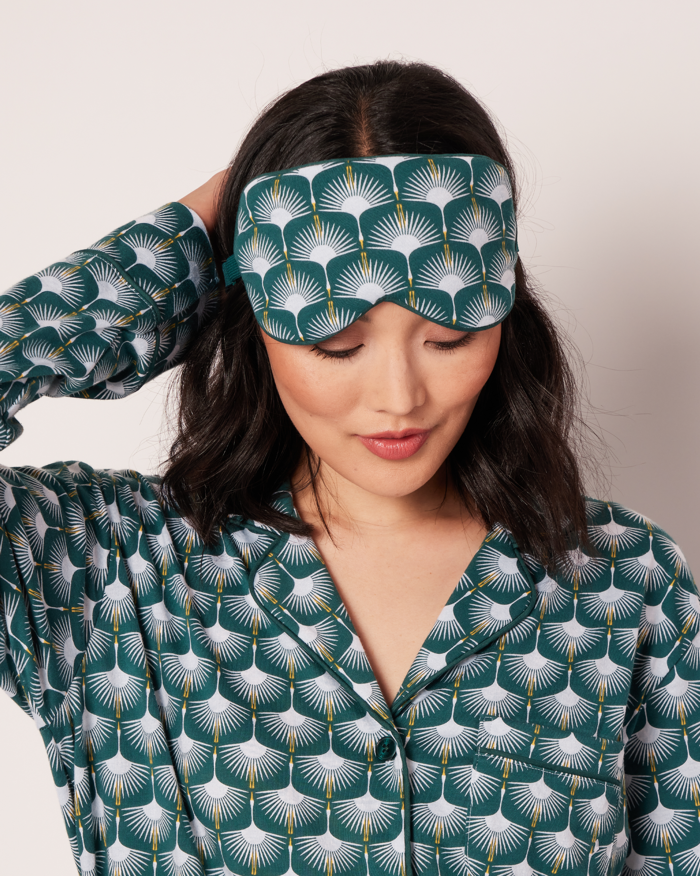 With dark hair, a person wears Petite Plume’s Womens Pima Sleep Mask in Sonnet of Swans and a matching pajama top, showcasing a teal and white fan design accented by swans. They glance down, emphasizing the elegant ensemble against a plain backdrop.