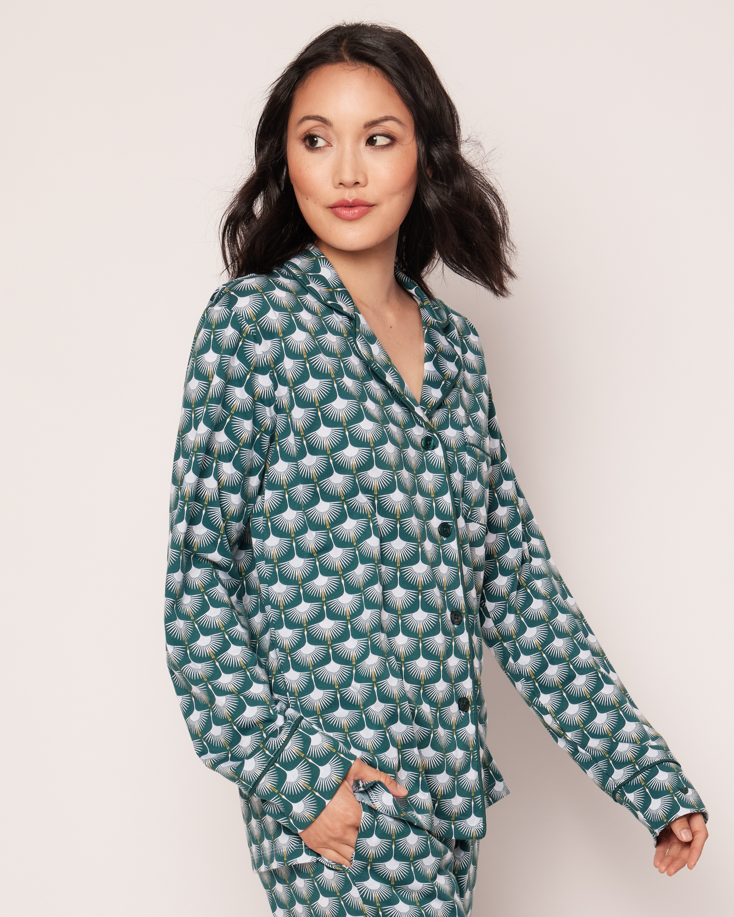 A woman with shoulder-length dark hair wears the Womens Pima Pajama Set in Sonnet of Swans by Petite Plume, showcasing green, blue, and white geometric swan designs. Made from luxurious Peruvian Pima cotton, she stands against a plain background, gazing over her left shoulder.
