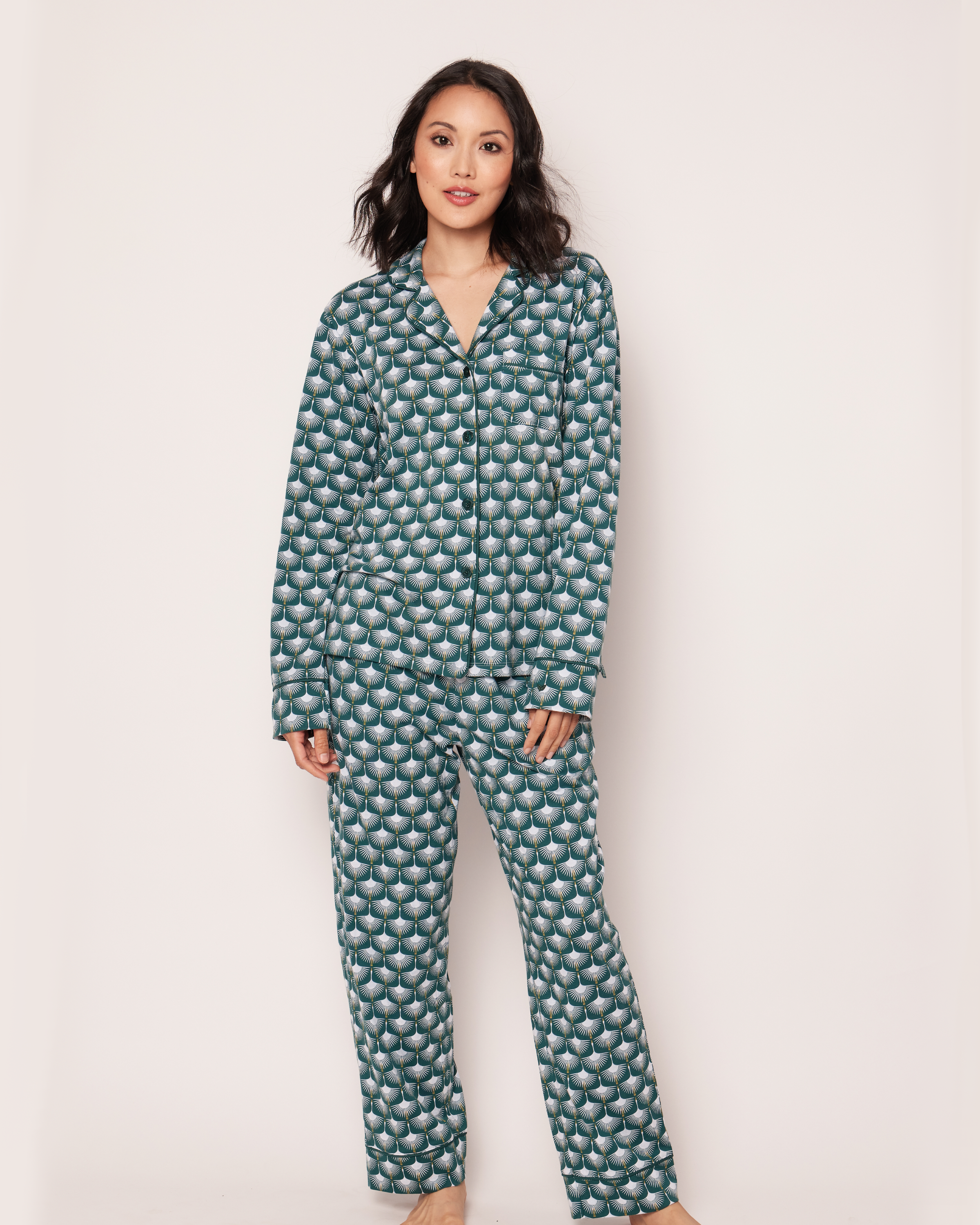 A person with shoulder-length dark hair in the Petite Plume Womens Pima Pajama Set, Sonnet of Swans, featuring a teal geometric swan motif, stands against a light background and looks towards the camera.