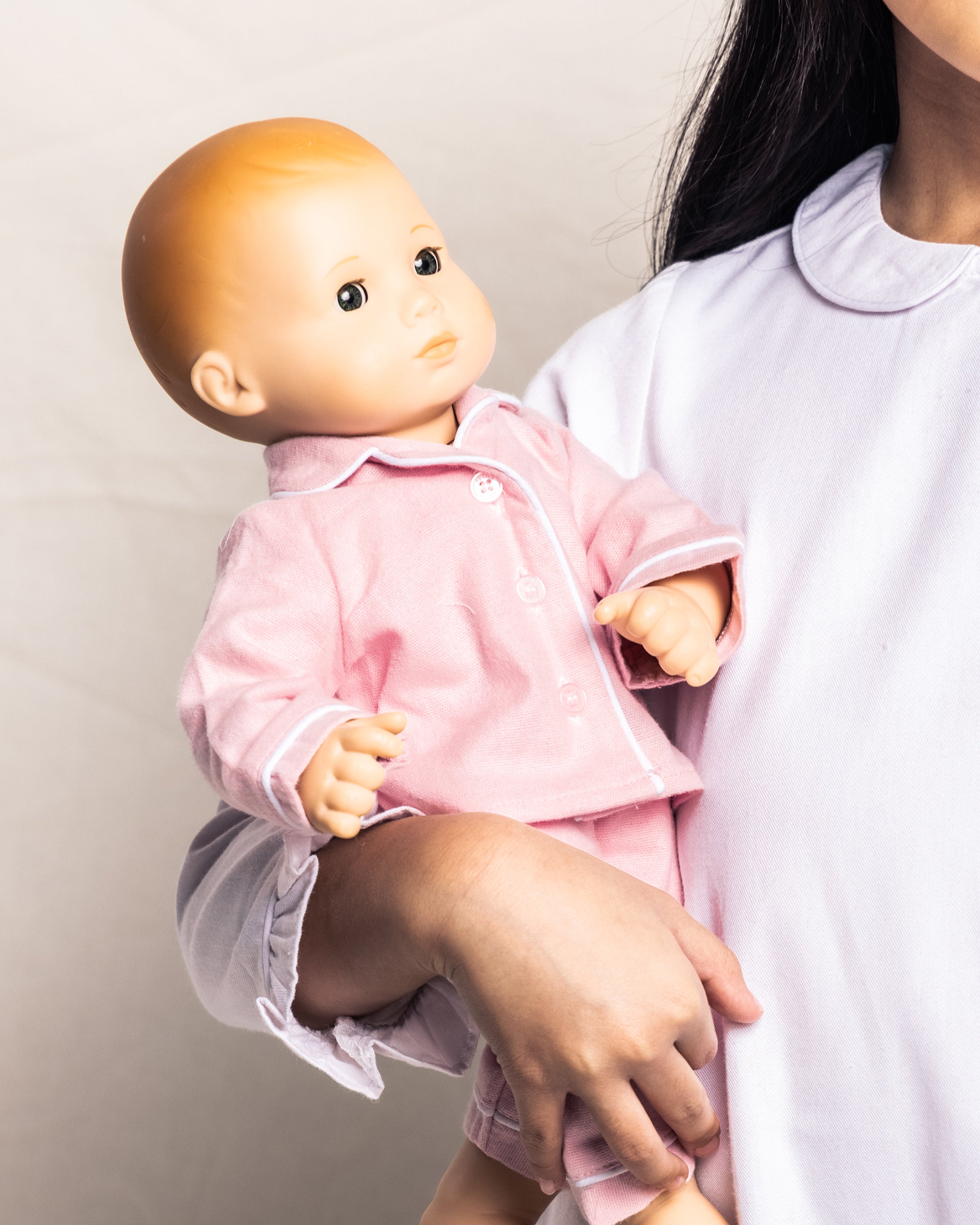A person in a long-sleeved white shirt holds an adorable 18 baby doll with a light skin tone, dressed in Petite Plumes Kids Flannel Doll Pajamas in Pink, perfectly matching their own attire. Only the shoulder and one arm of the individual are visible.