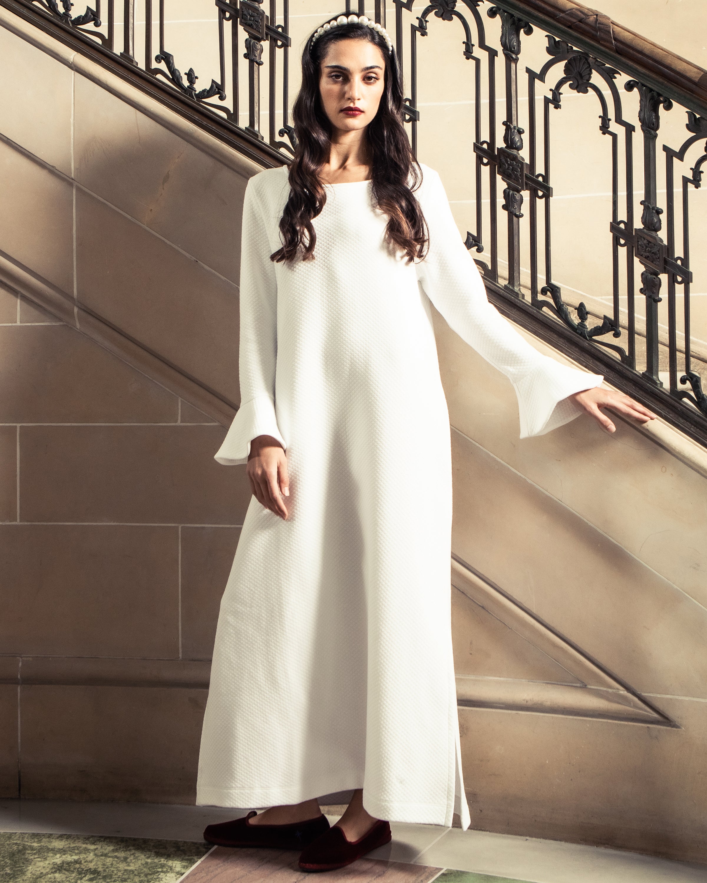 A woman in a long, white dress with bell sleeves stands by a staircase, wearing Petite Plumes Womens Hamptons Loafer in Bordeaux Velvet and a headband in her long dark hair. The stair railing features ornate ironwork, enhancing the elegant classical setting.