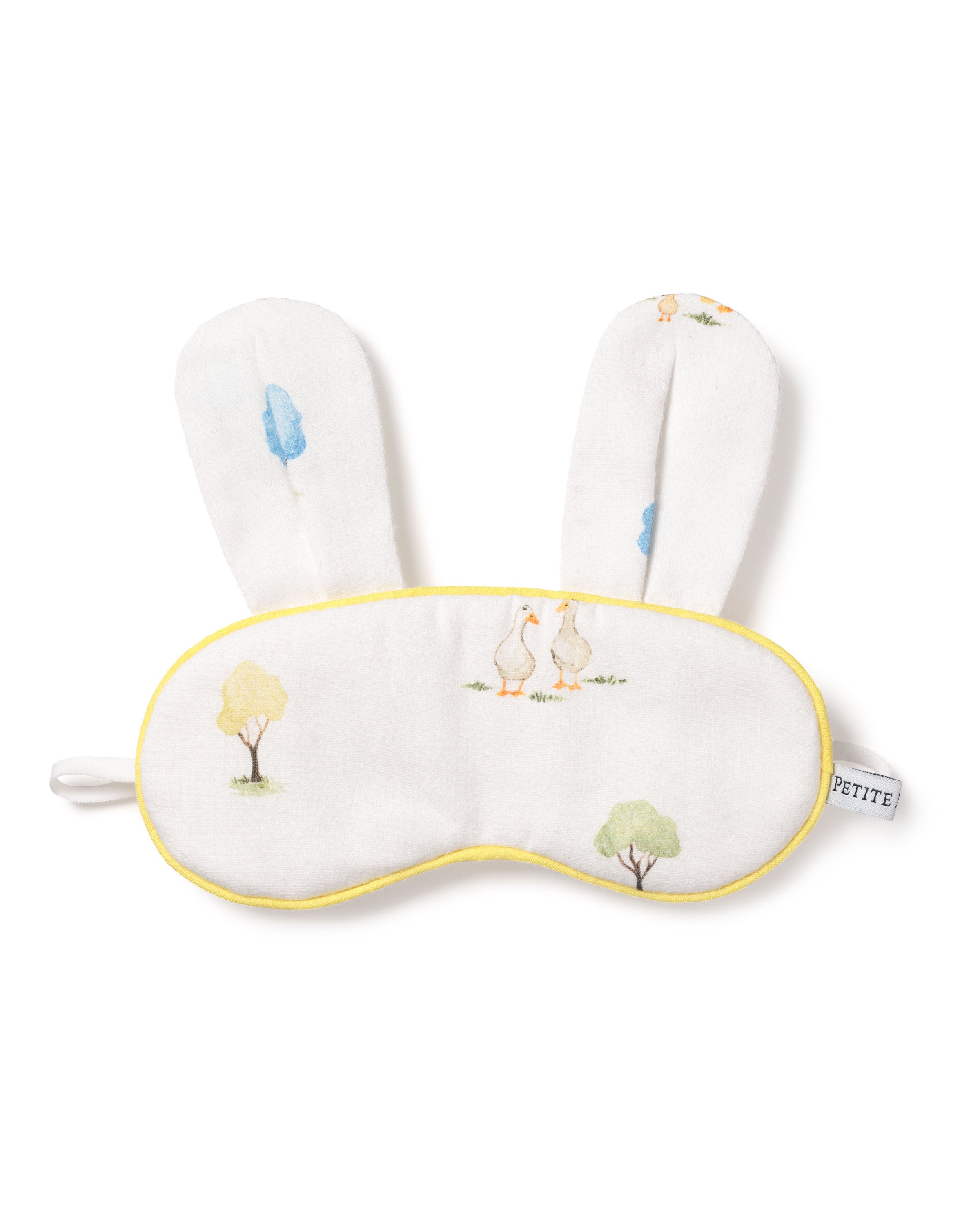 The Kids Twill Bunny Sleep Mask in Easter Gardens by Petite Plume is a luxurious soft white mask featuring yellow trim, bunny ears, whimsical tree illustrations, and geese motifs. This charming piece is an ideal monogrammed gift with a small side tag labeled PETITE.