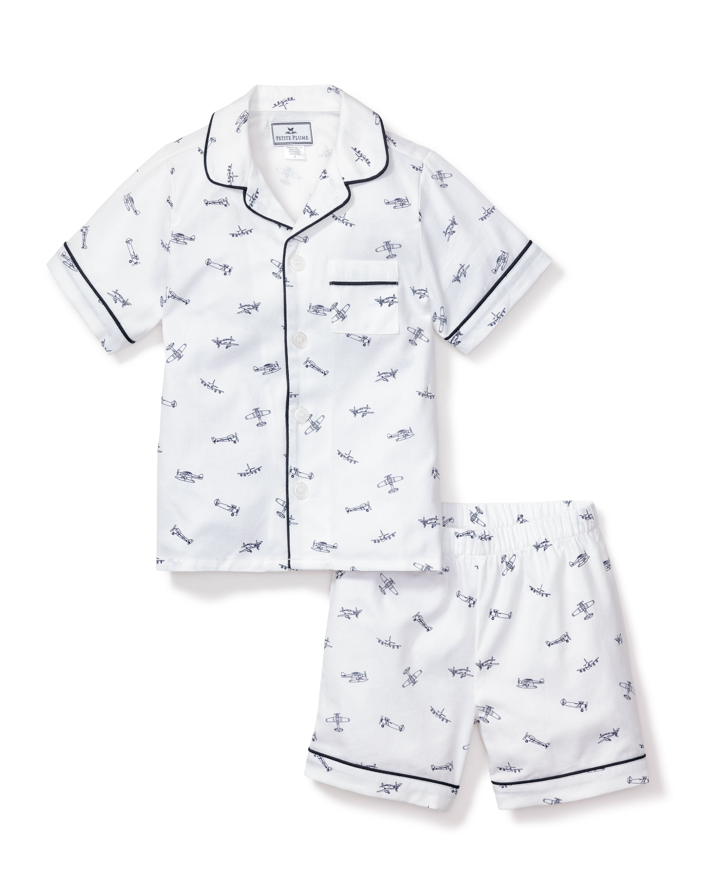 The Petite Plume Kids Twill Pajama Short Set in Par Avion offers luxurious sleepwear with a white short-sleeve button-up shirt featuring navy piping, a chest pocket, and playful prints of planes, helicopters, and vehicles, paired with matching shorts.
