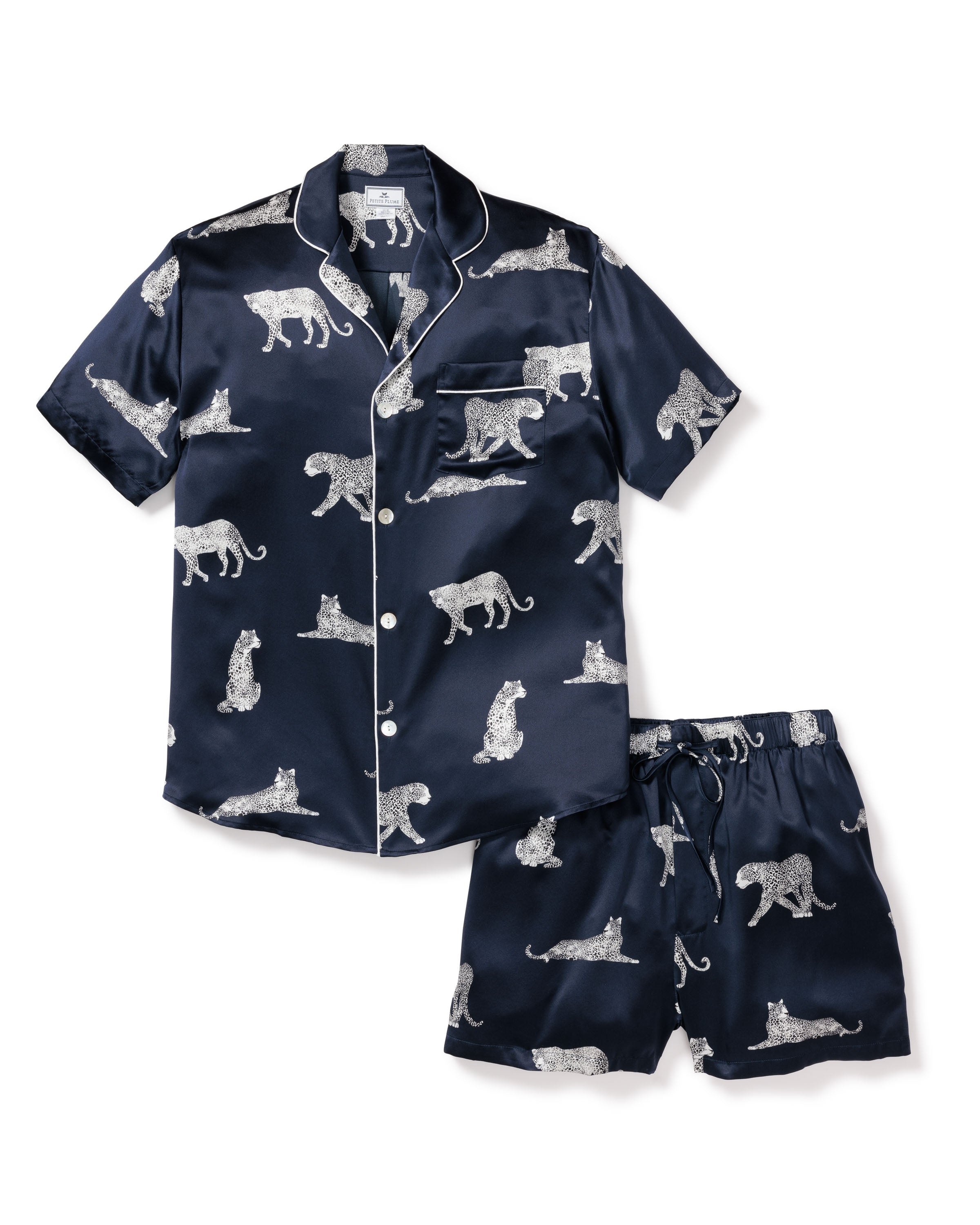 The Mens Silk Pajama Short Set in Panthère de Nuit by Petite Plume features navy Mulberry silk with short sleeves and shorts, accented by white piping. Hypoallergenic fabric decorated with white leopards ensures a stylish and comfy nights rest.