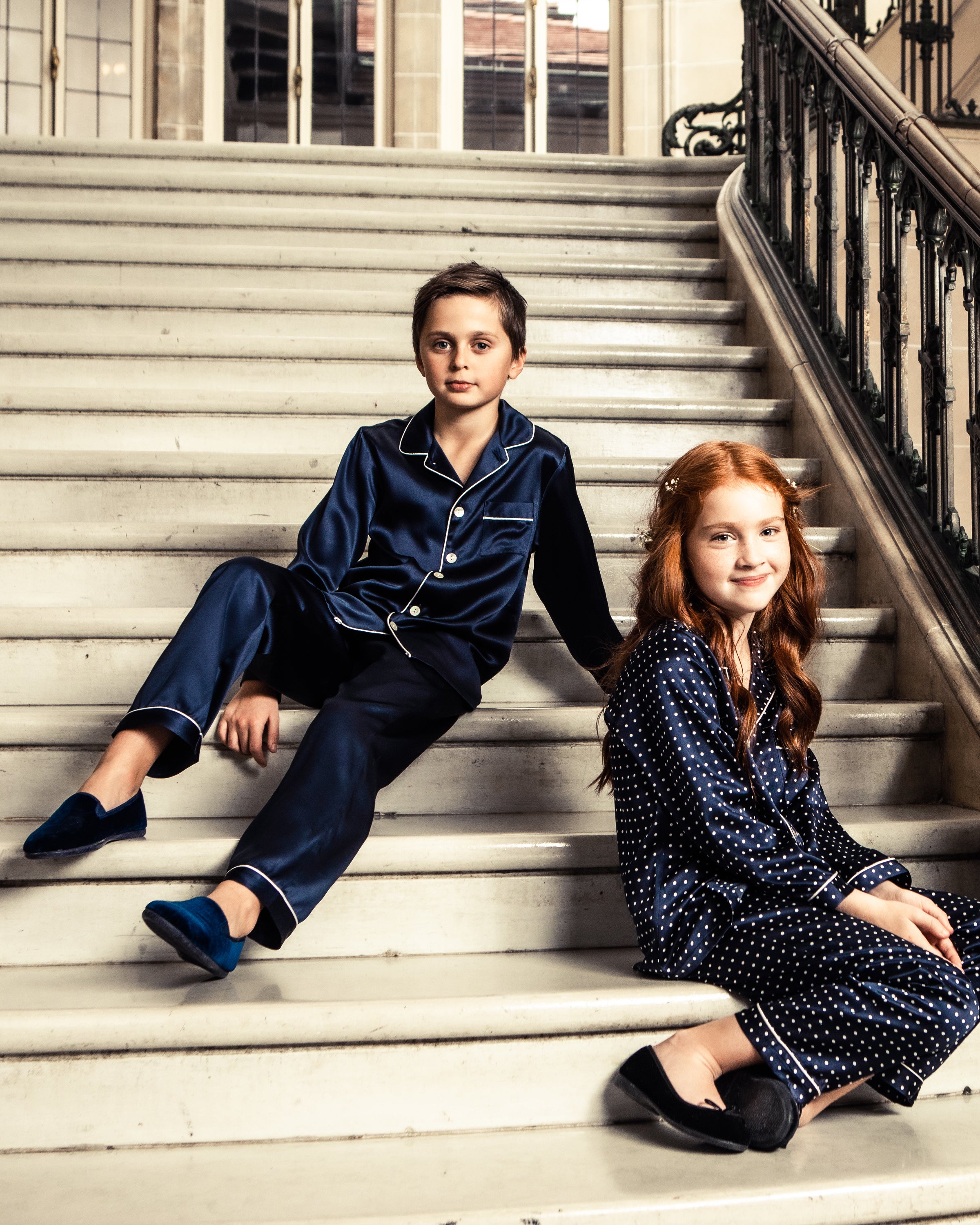 Two children sit on an elegant staircase with ornate railings in a grand building—one with short hair in warm sleepwear and Kids Gable Slipper in Navy Velvet by Petite Plume, and the other with long red hair wearing polka-dotted pajamas.