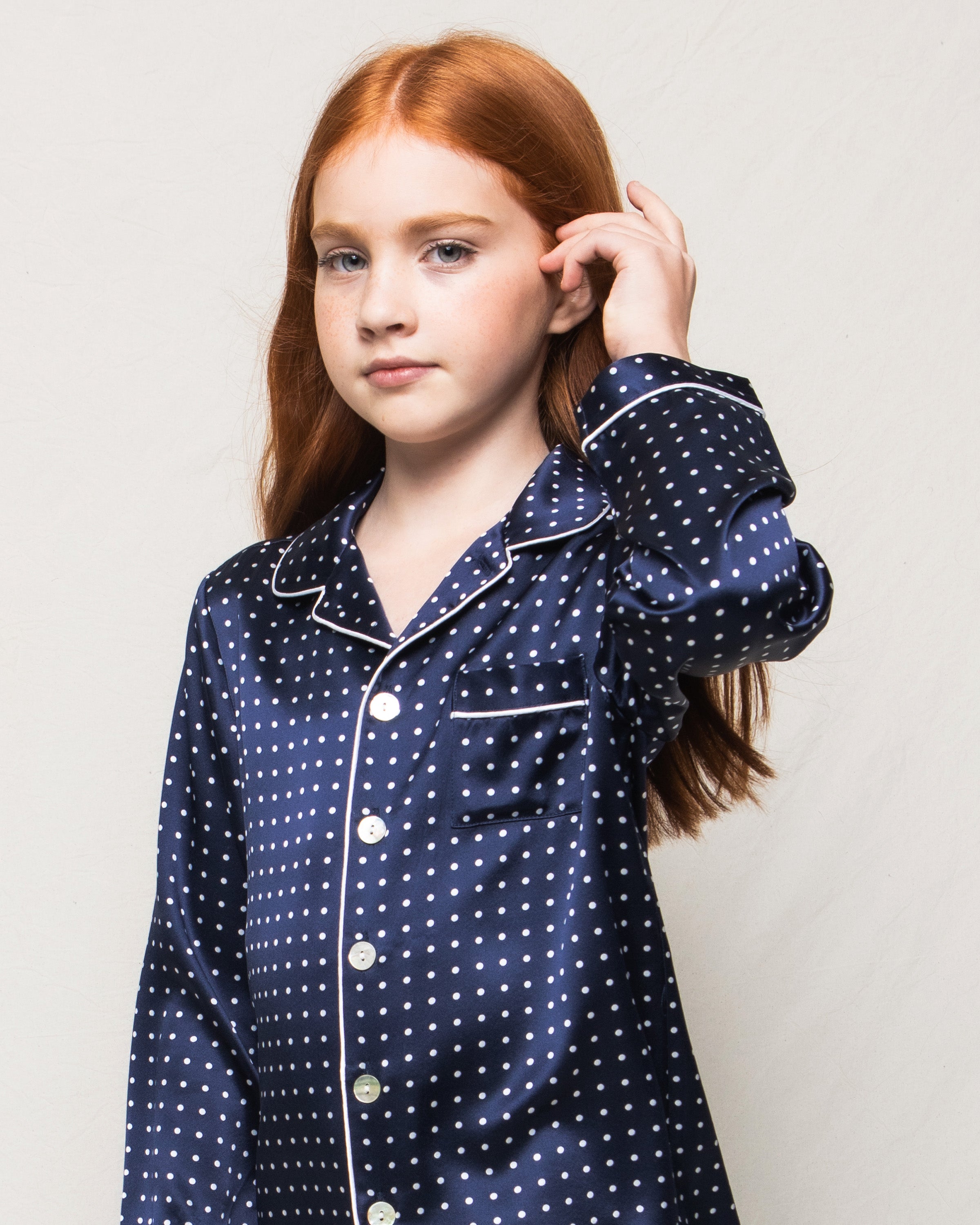 A young girl with long red hair wears the Petite Plume Kids Silk Pajama Set in Navy Polka Dot. She gazes at the camera, gently brushing her hair back with one hand against a light-colored, plain background.