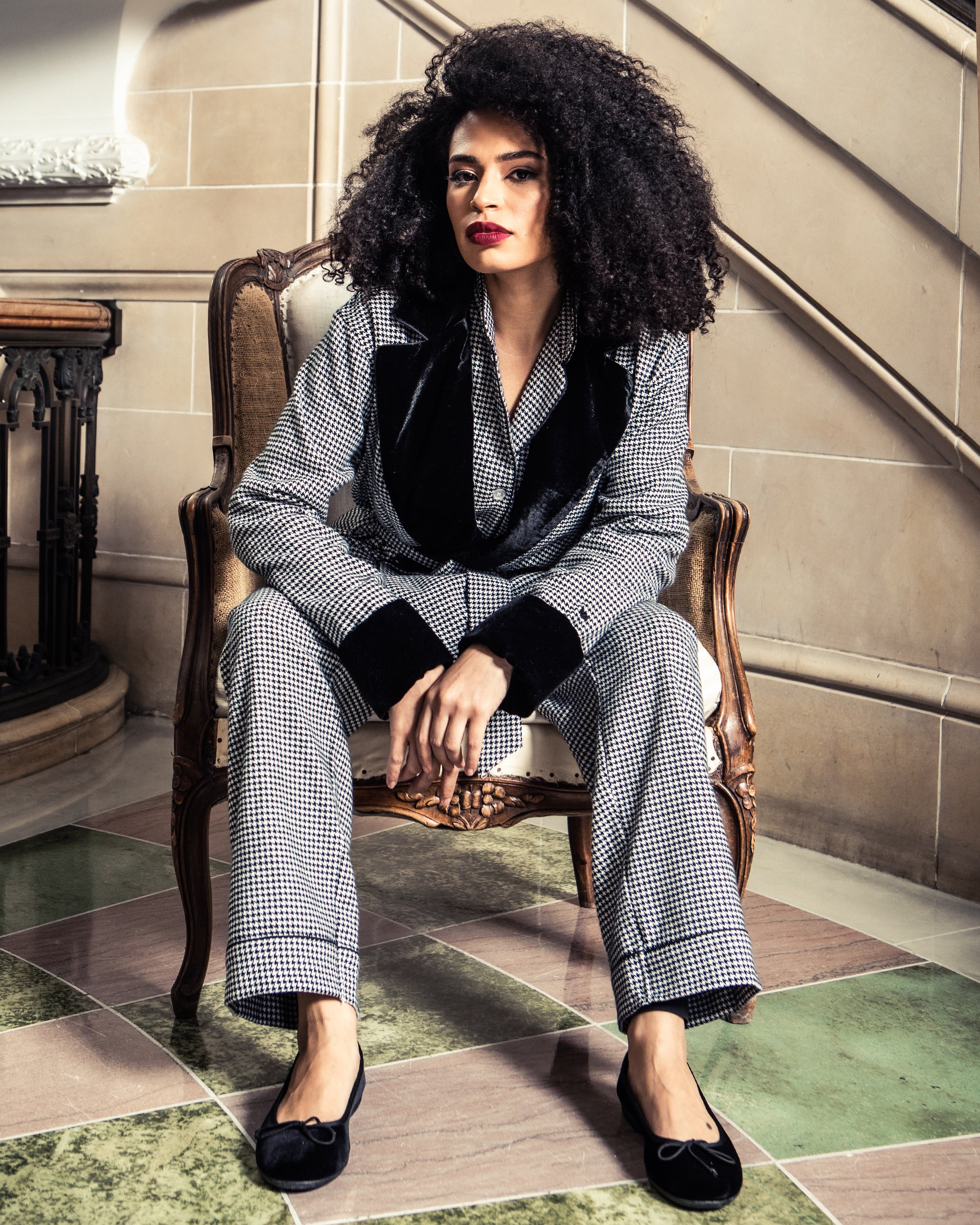 A person with voluminous curly hair sits confidently in a vintage armchair, wearing a chic houndstooth suit with a luxe velvet collar and Petite Plumes Womens Juliette Ballet Slipper in Black Velvet. The scene is set against elegant stone walls and a decorative bannister.
